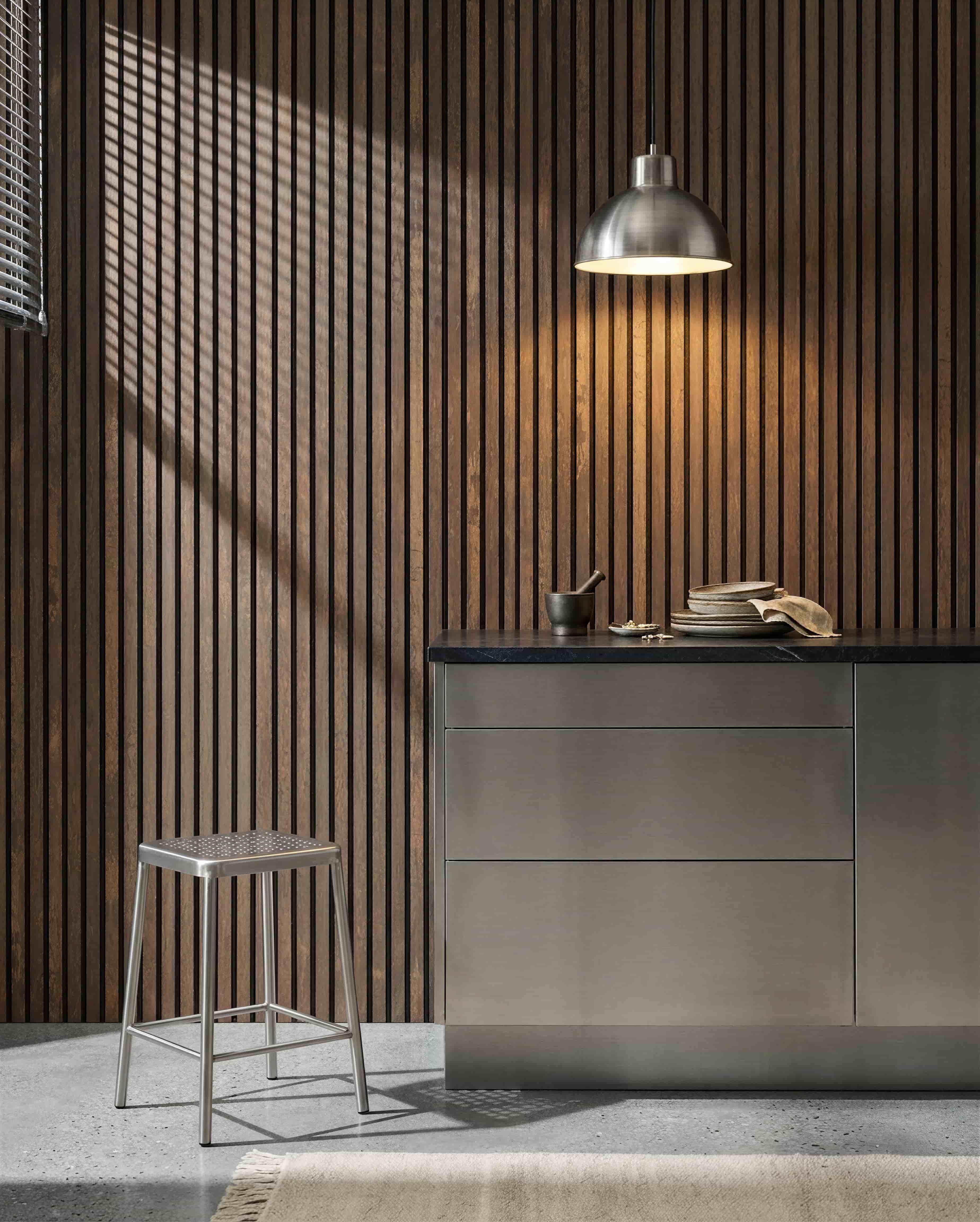 Kuchnia z akustyczną ścianą lamelową WoodUpp Akupanel Oxide Copper, blatem ze stali nierdzewnej, metalowym stołkiem, lampą wiszącą, różnymi naczyniami oraz moździerzem i tłuczkiem.