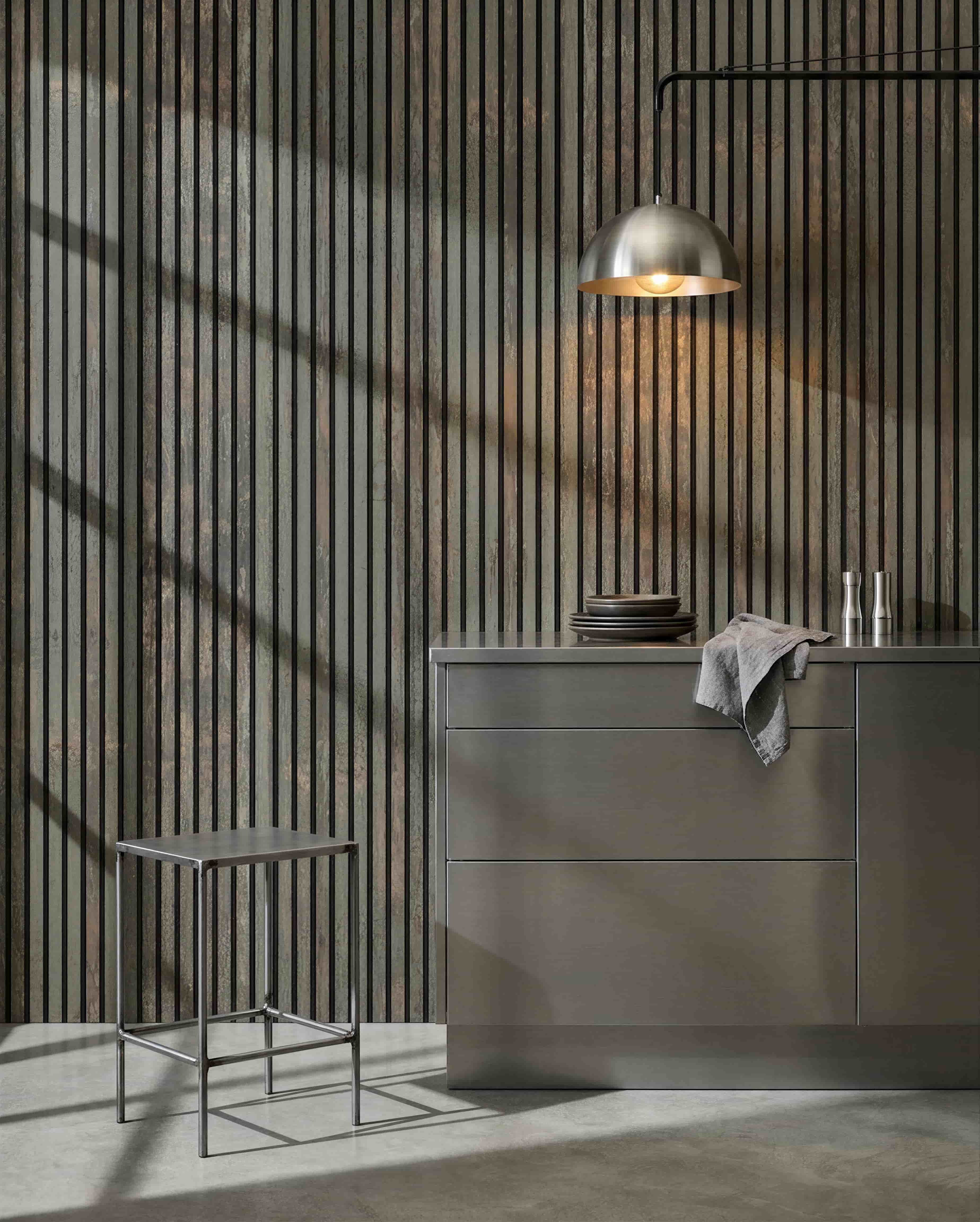 Minimalistyczna kuchnia z metalowym stołkiem, wyspą grey i naczyniami, z akustycznym panelem drewnianym WoodUpp Akupanel w kolorze Oxide Grey/Black Felt, który pomaga kontrolować dźwięk.