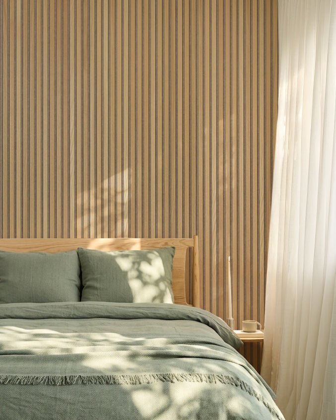 Minimalistyczna nordycka sypialnia z dębową ścianą WoodUpp Akupanel Panel akustyczny, pościelą w kolorze szałwiowej zieleni, drewnianą ramą łóżka i lekkimi białymi zasłonami.