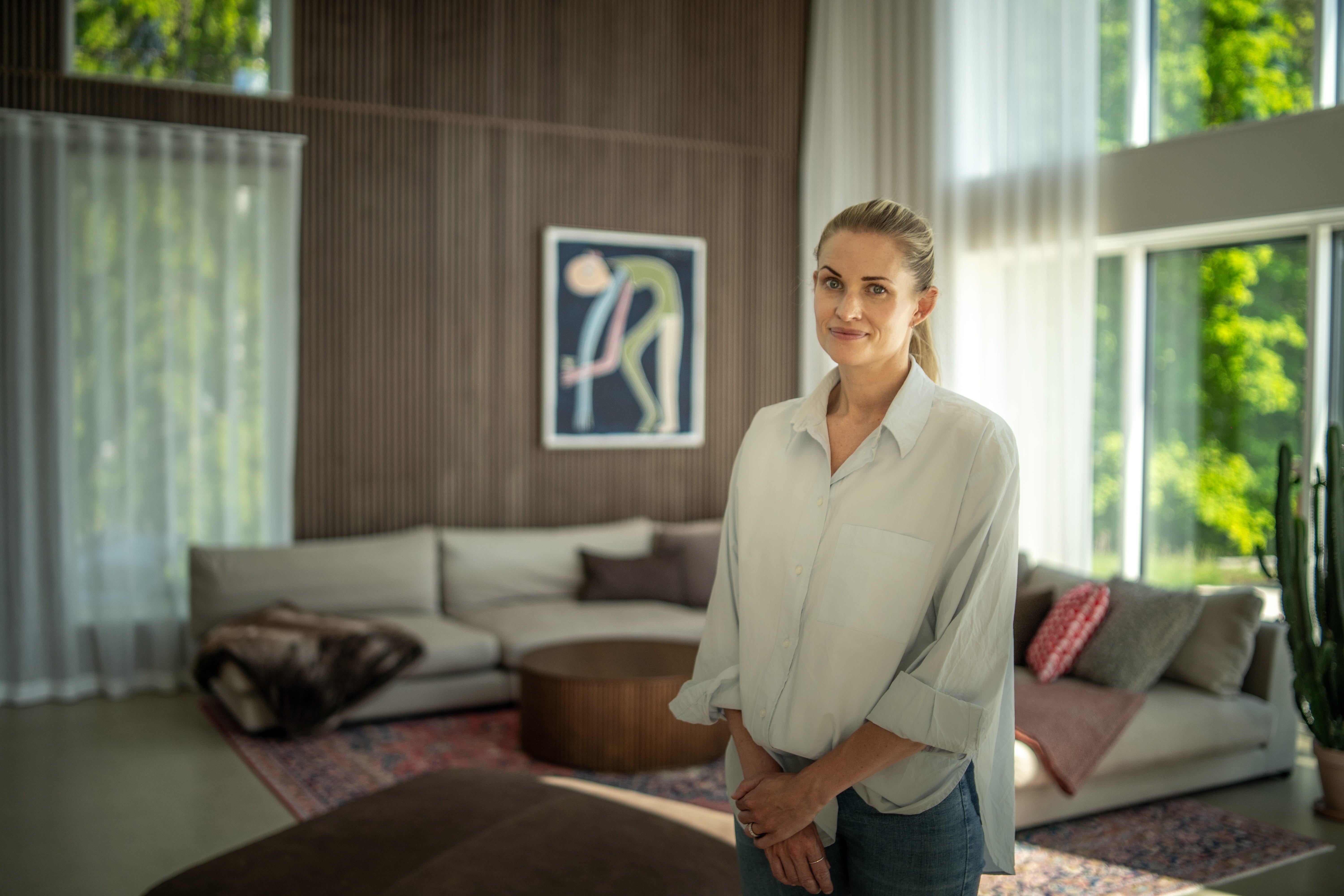 Kobieta stoi w nasłonecznionym salonie z dużymi oknami, skandynawskimi panelami z drewna akustycznego na ścianie, sztuką współczesną i neutralnymi, minimalistycznymi meblami.