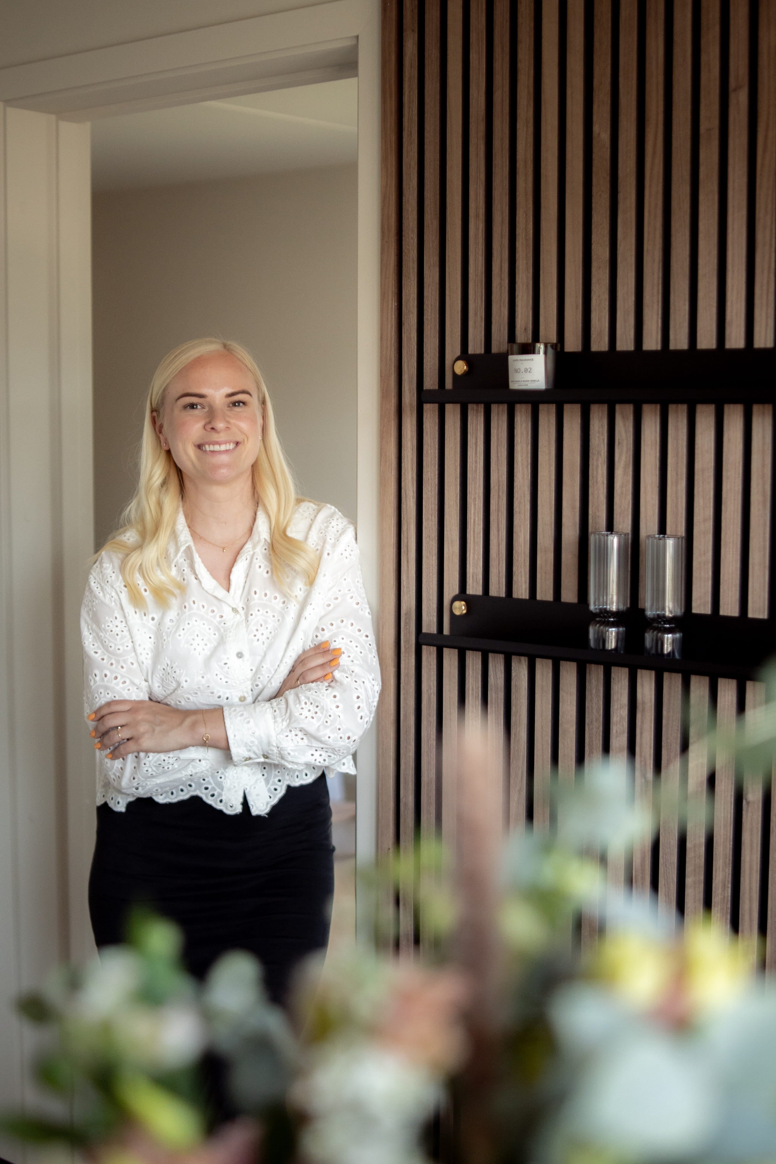 Kobieta z długimi blond włosami i białą bluzką stoi uśmiechnięta ze skrzyżowanymi ramionami w nowoczesnym pokoju z panelami z drewna akustycznego i eleganckimi półkami.