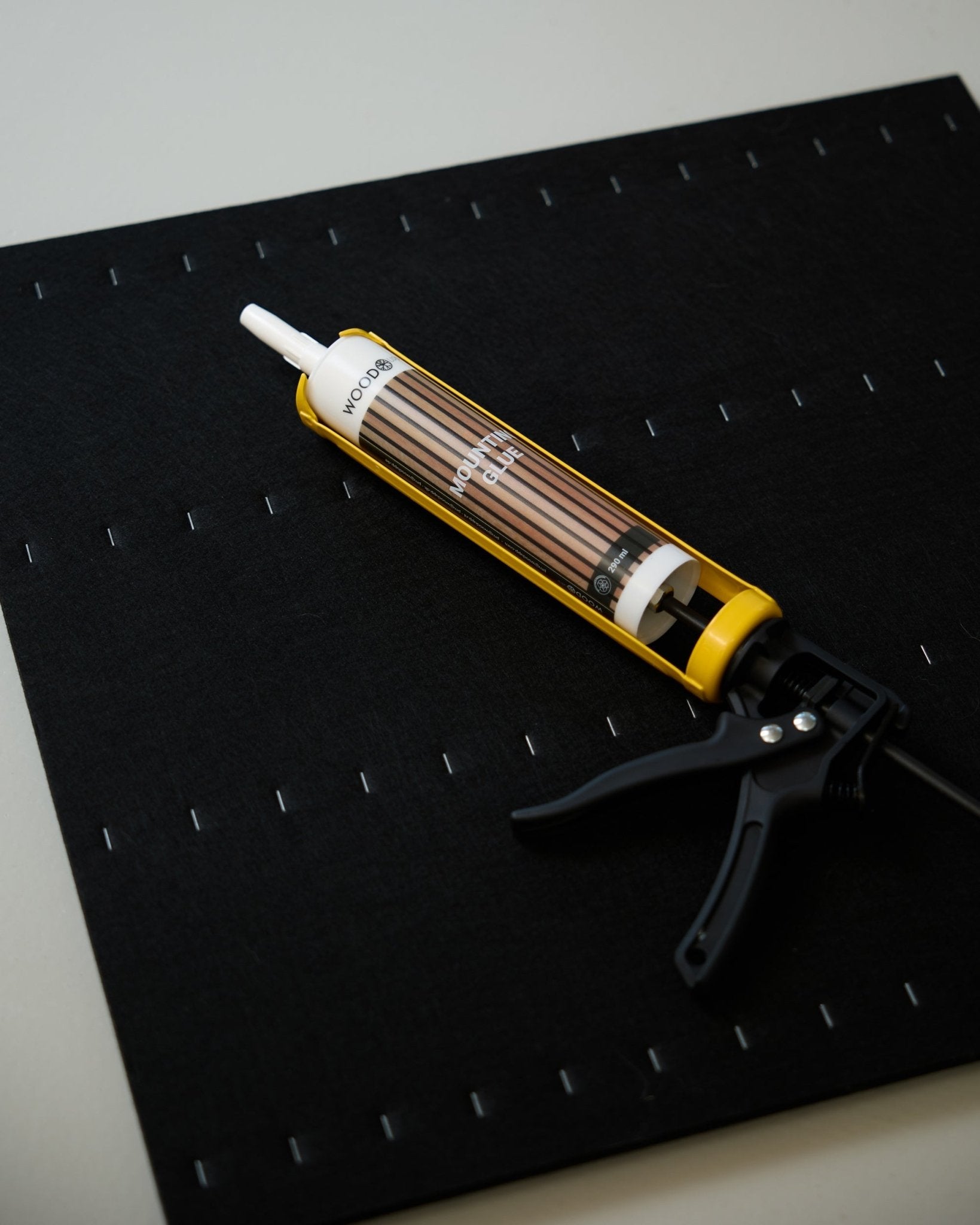 Żółty i black WoodUpp Grout Gun (290 ml) leży na black macie z siatką białych zszywek i służy do montażu paneli akustycznych.