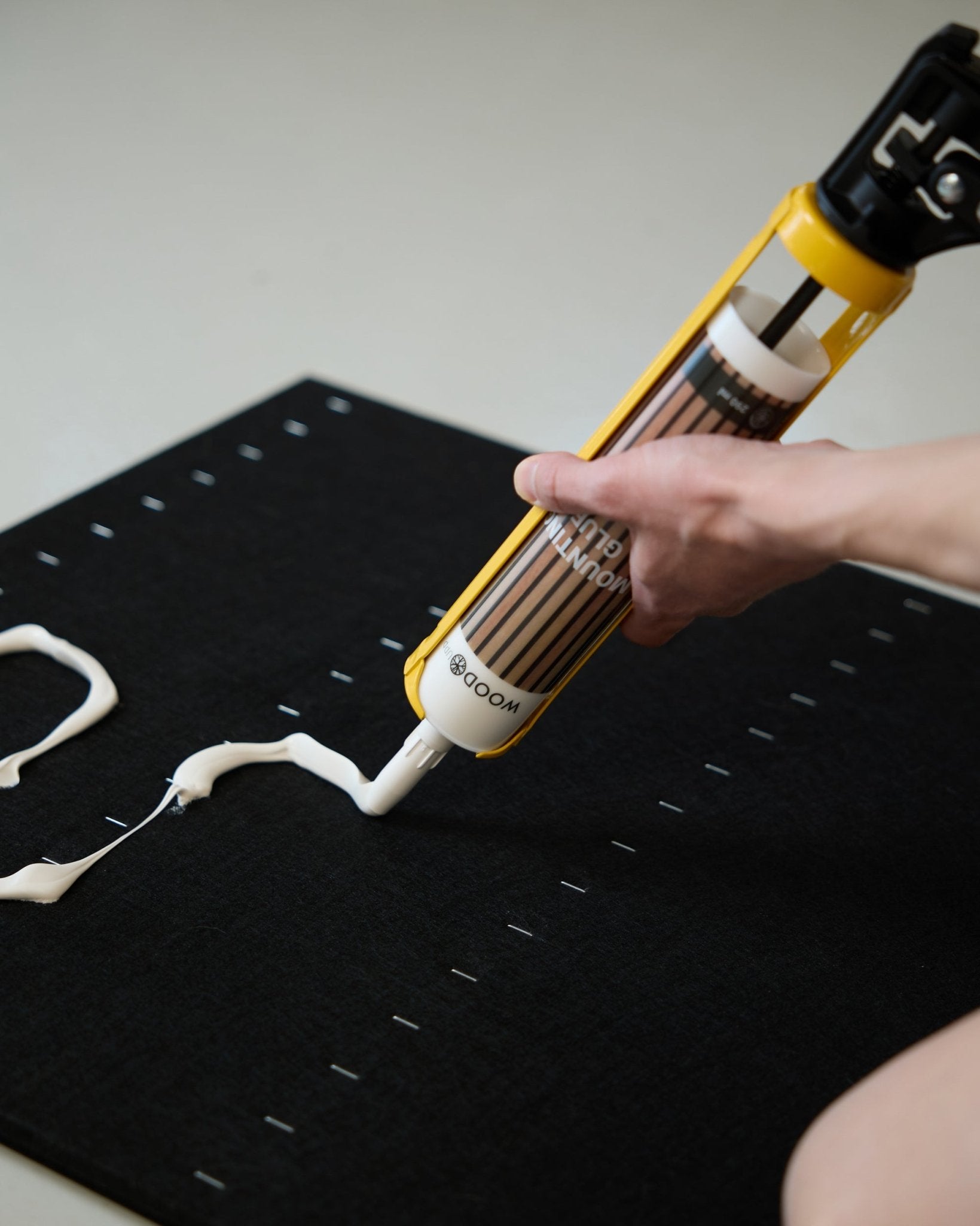 Dłoń nakłada biały klej montażowy zygzakowatym wzorem za pomocą WoodUpp Grout Gun (290 ml) na powierzchnię black Panel akustyczny.