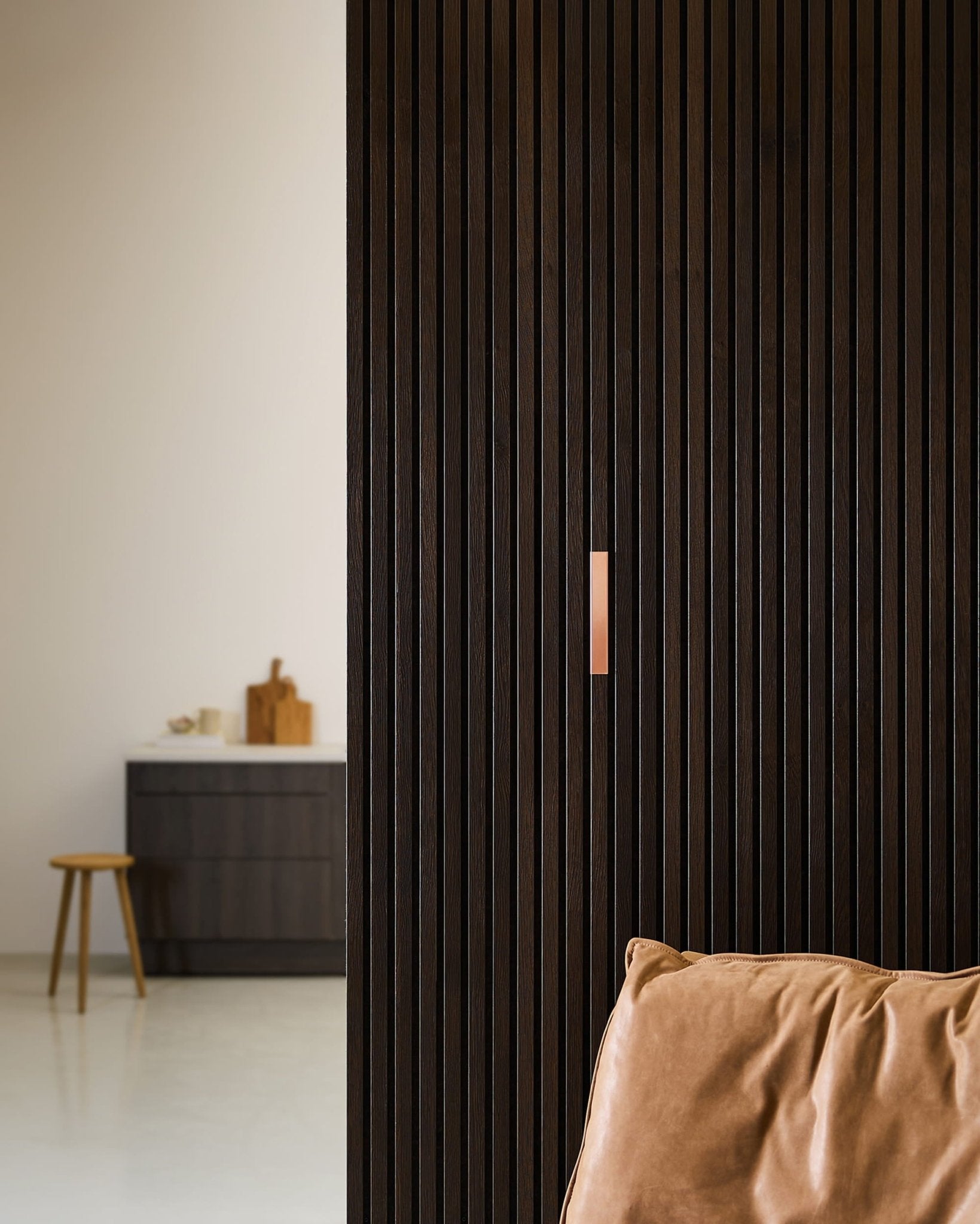 Minimalistyczne wnętrze z brązowym skórzanym fotelem WoodUpp Liva | Cover / Copper, 20-centymetrowymi panelami akustycznymi na ścianie oraz stołkiem przy blacie z deskami do krojenia.