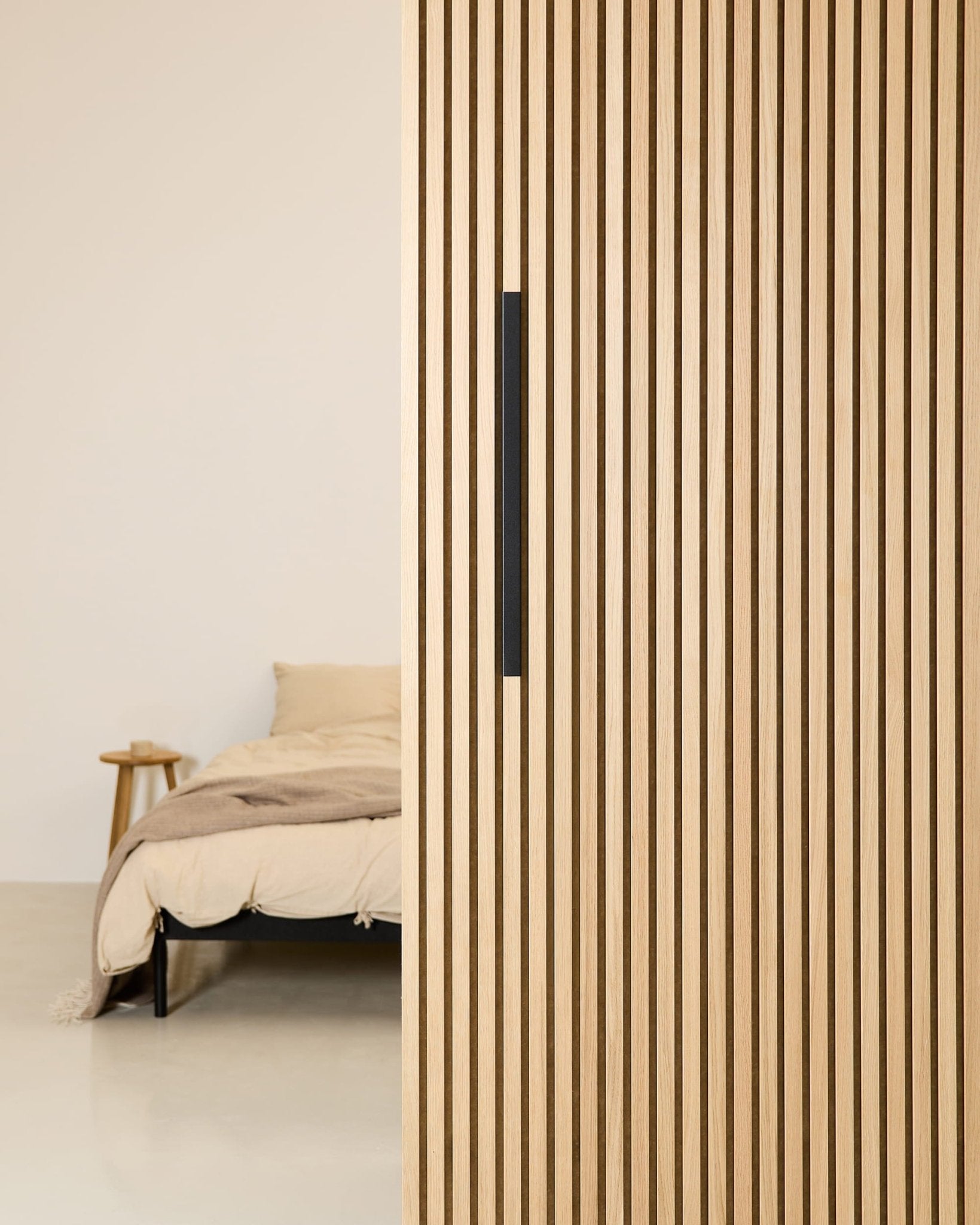 Minimalistyczna sypialnia z beżowym łóżkiem, częściowo zasłoniętym przez czarną aluminiową przegrodę WoodUpp EMBRACE o szerokości 60 cm, wyposażoną w czarny uchwyt.