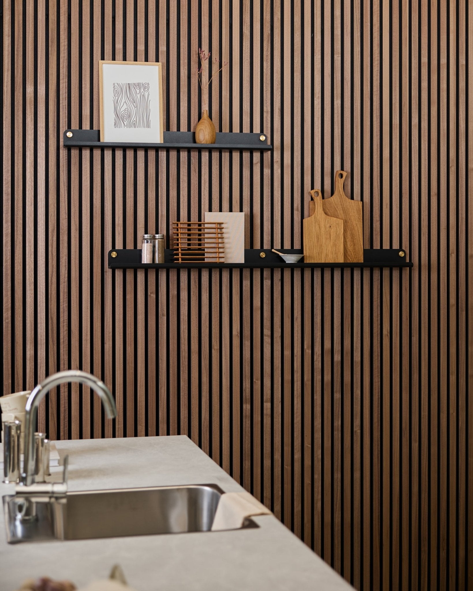 Minimalistyczna kuchnia z akustyczną ścianą z drewnianych lameli w kolorze vertical Akupanel, półkami black, deskami do krojenia, dekoracjami small oraz nowoczesnym blatem ze zlewem na pierwszym planie.