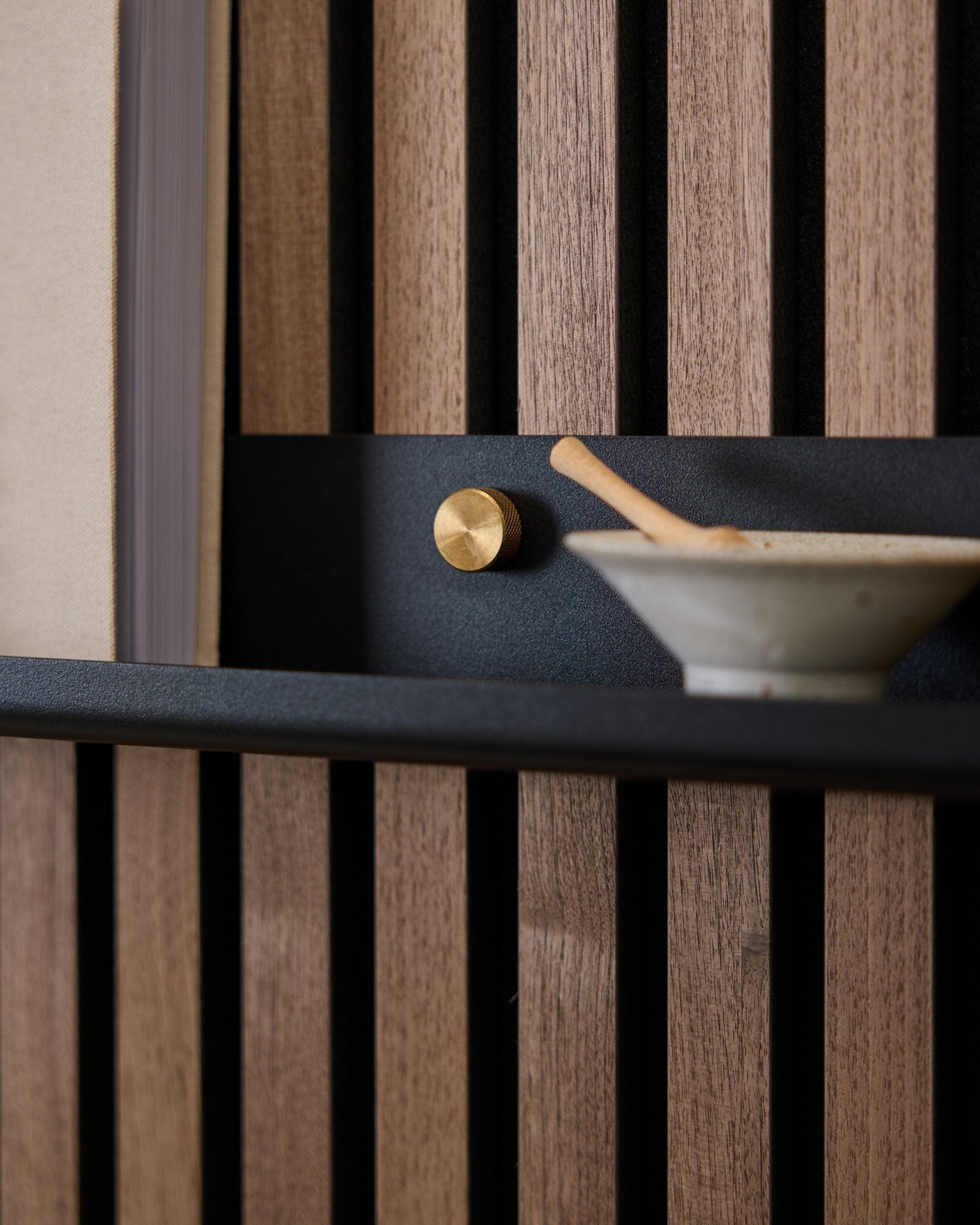 Zbliżenie na półkę Narrow Shelf WoodUpp Norr o długości 115,9 cm w kolorze black, zamontowaną na nordyckim akustycznym panelu lamelowym za pomocą śrub radełkowanych brass, ze złotym pokrętłem oraz ceramiczną miską z łyżką.