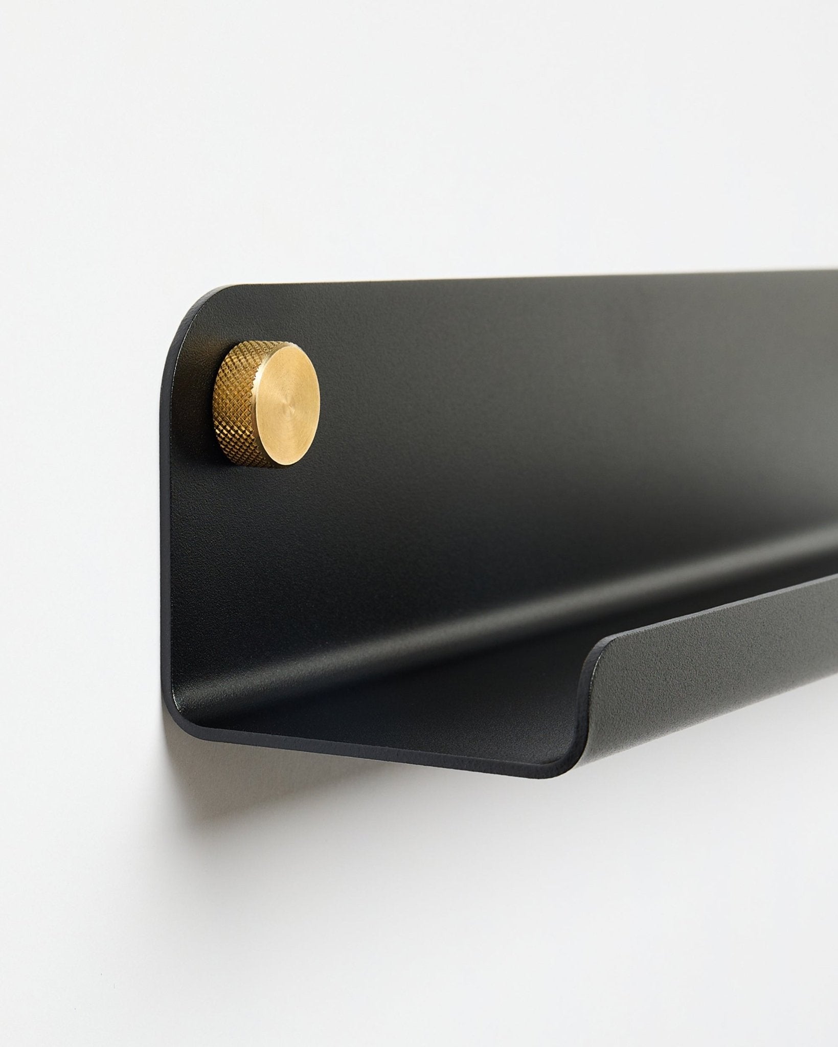 Norr | Półka Narrow Shelf w kolorze black (115,9 cm) z jednym złotym pokrętłem po lewej stronie, od WoodUpp, zamontowana na jednolitej białej ścianie.