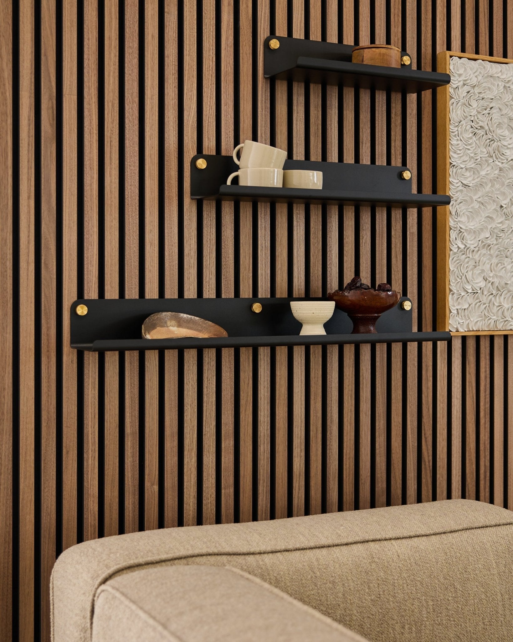 Trzy półki w kolorze black, w tym szeroka półka WoodUpp Norr Wide Shelf (75,9 cm), eksponują dekoracje na ścianie z lameli Panel akustyczny nad sofą w kolorze beige; fragment obrazu jest widoczny.