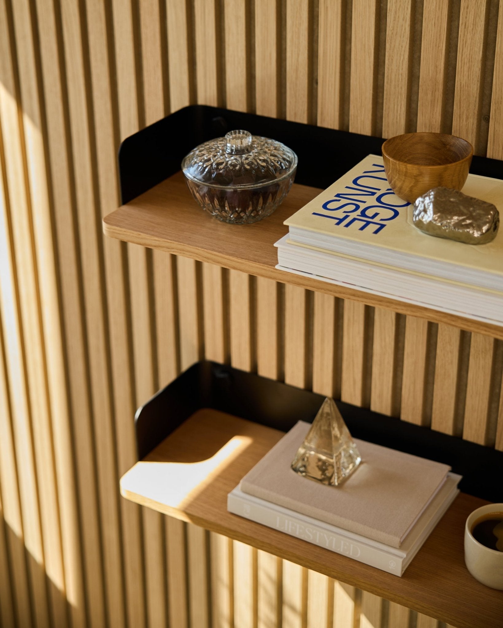 Biblioteczka WoodUpp Linn w kolorze Natural Oak (55,9 cm) mieści szklany słoik, filiżankę, złotą piramidę i książki na tle akustycznych paneli drewnianych Akupanel w promieniach słońca.