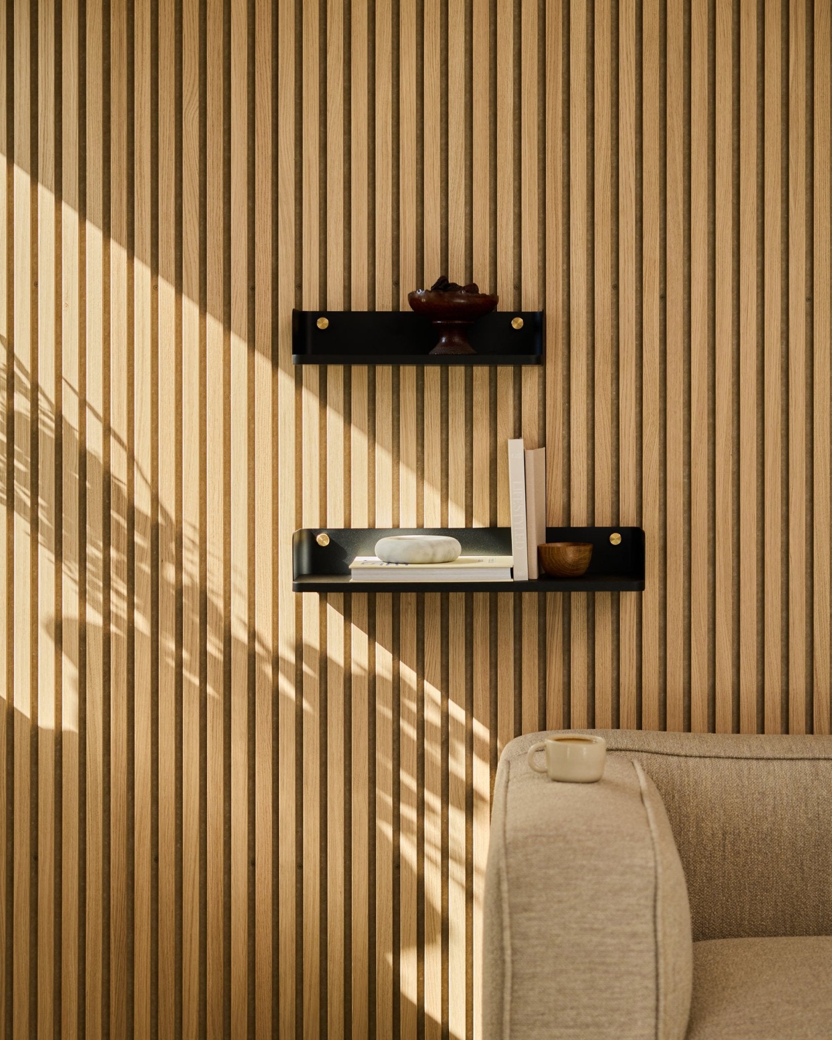 Światło słoneczne oświetla sofę beige oraz półkę na książki Linn marki WoodUpp w kolorze black oak, 55,9 cm, na tle minimalistycznego nordyckiego panelu ściennego z listew Panel akustyczny.