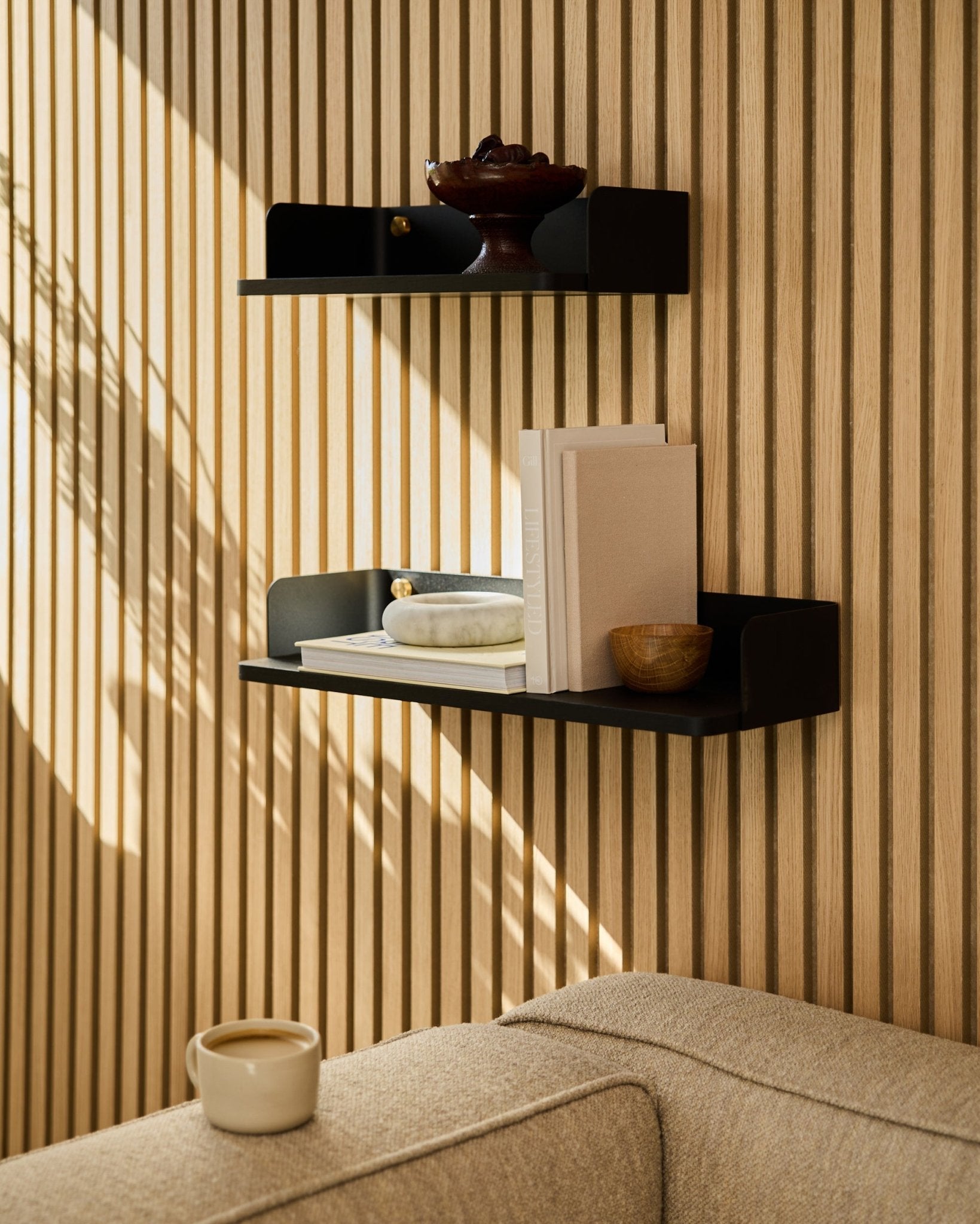 Półka na książki WoodUpp black oak (55,9 cm) eksponuje książki, dekoracje i miskę na ścianie z akustycznych paneli drewnianych; na pierwszym planie sofa beige i kubek.