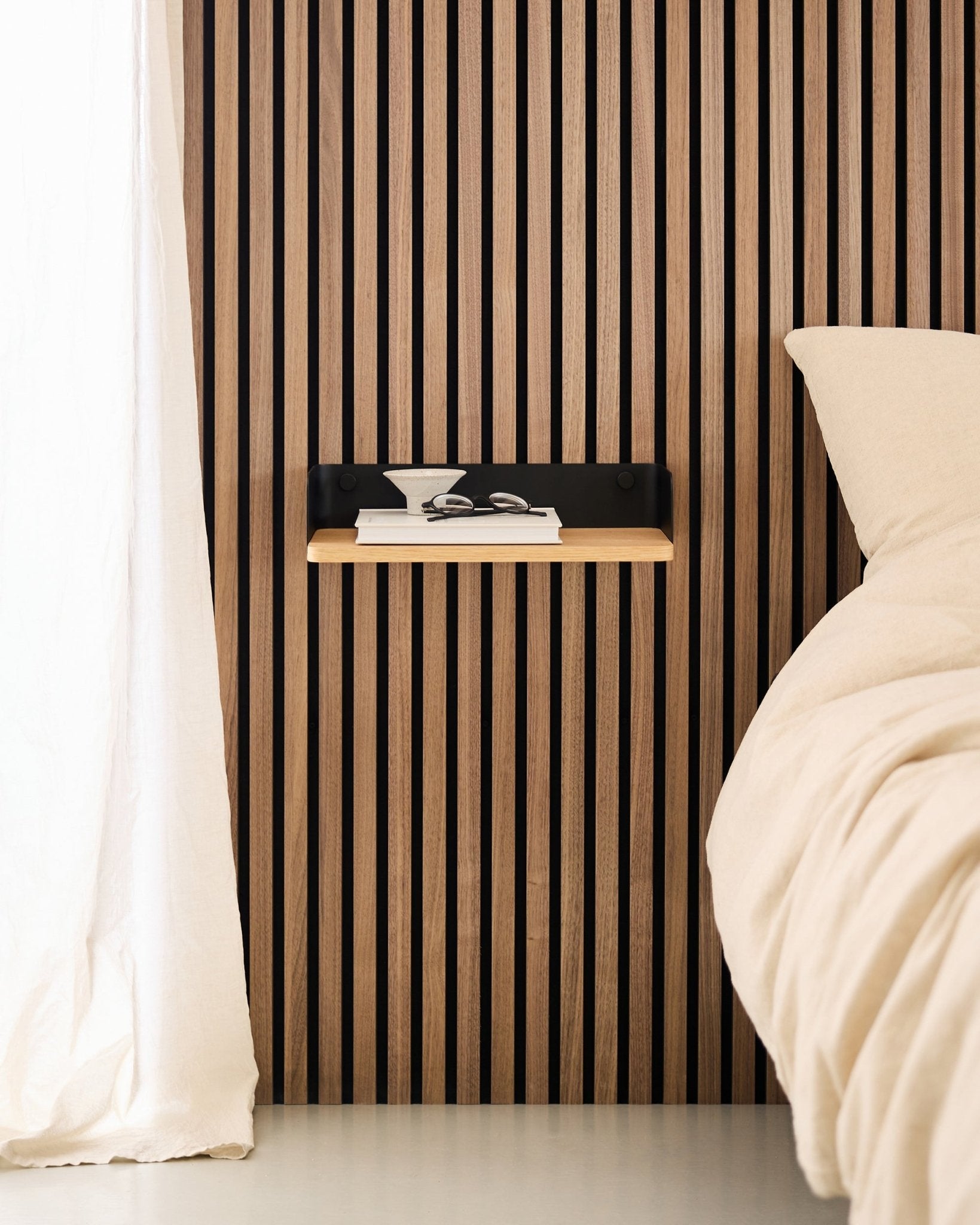 Minimalistyczna sypialnia ze ścianą z nordyckich drewnianych lameli Panel akustyczny, półką na książki WoodUpp Linn w kolorze Natural Oak i wiszącym stolikiem nocnym obok łóżka beige.
