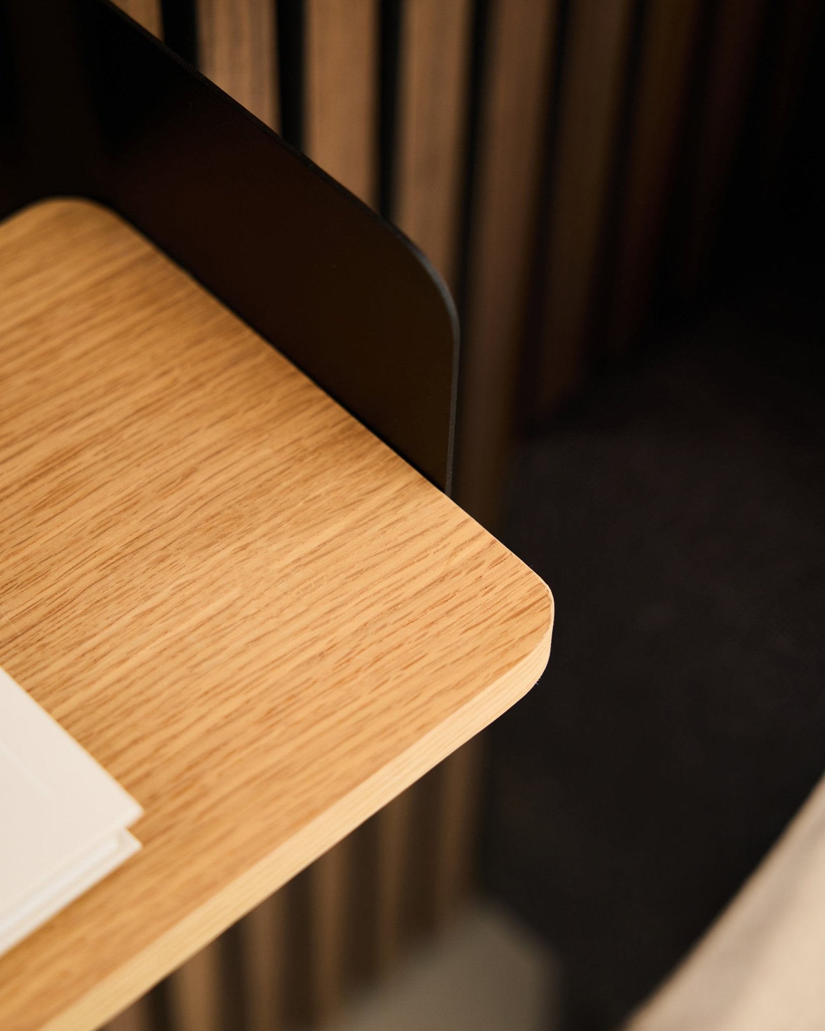 Zbliżenie na róg biurka z akustycznym panelowaniem drewnianym black Akupanel, białymi kartkami oraz półką na książki WoodUpp Linn w kolorze Natural Oak (39,9 cm) w tle.