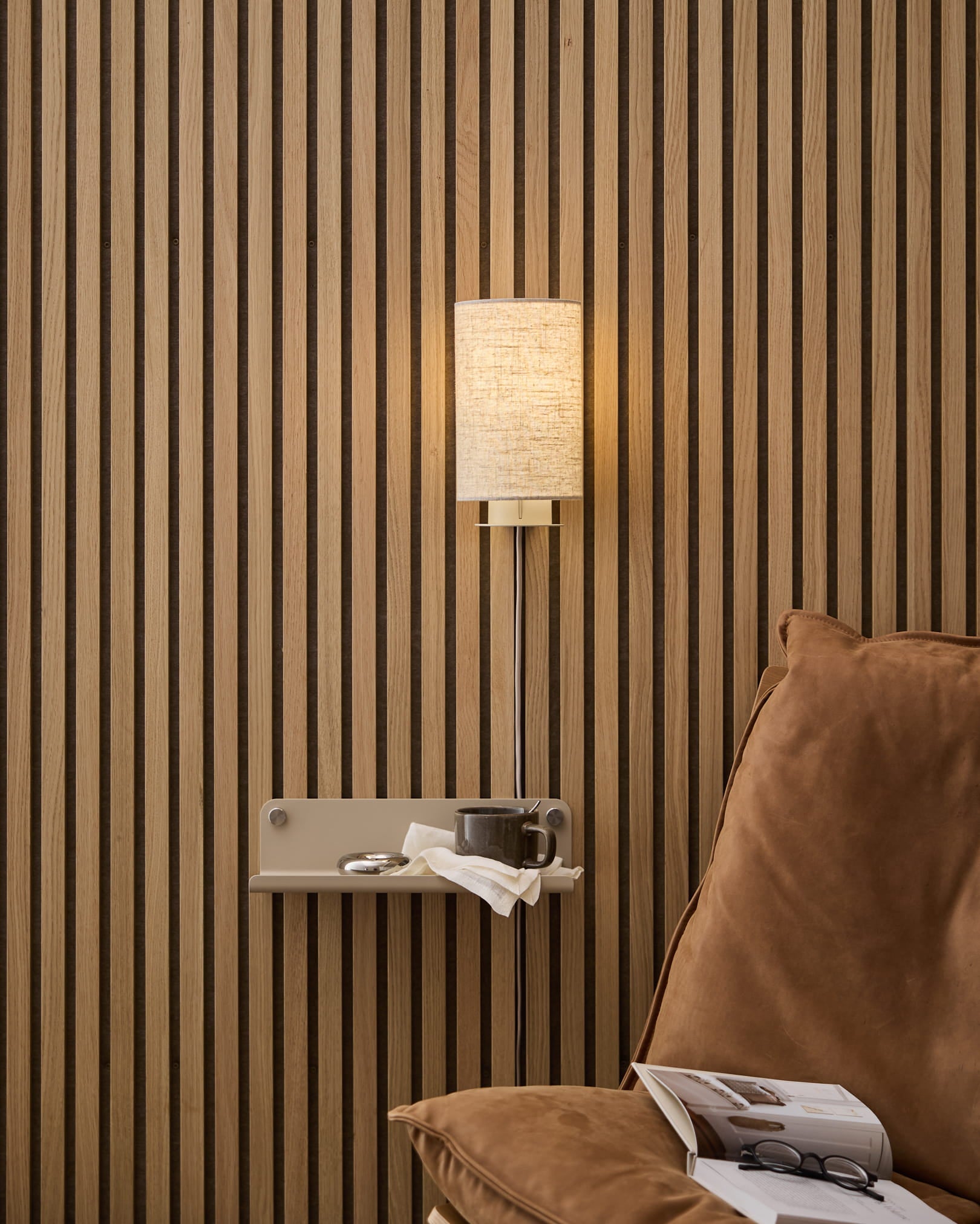 Lampa ścienna WoodUpp Feya w kolorze beige, z abażurem z lnu i bawełny, zamontowana nad przytulnym fotelem i stolikiem bocznym z książką oraz filiżanką, na tle nordyckiego drewnianego panelu lamelowego Panel akustyczny.