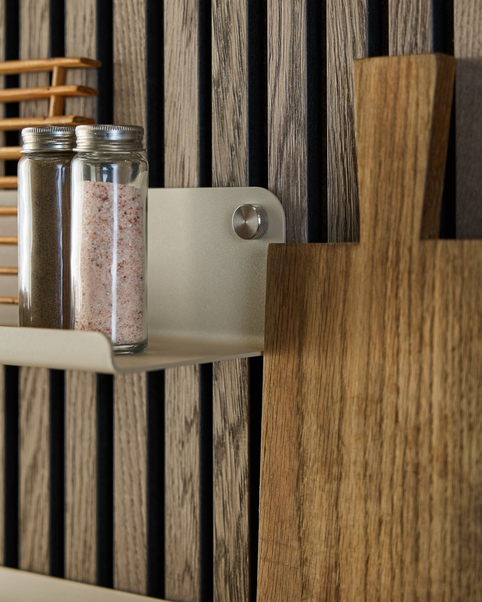 Na szerokiej półce WoodUpp Norr w kolorze beige, 35,9 cm, stoją szklane słoiki z przyprawami i drewniana deska do krojenia; półka jest zamontowana na akustycznym panelu lamelowym w kolorze vertical.