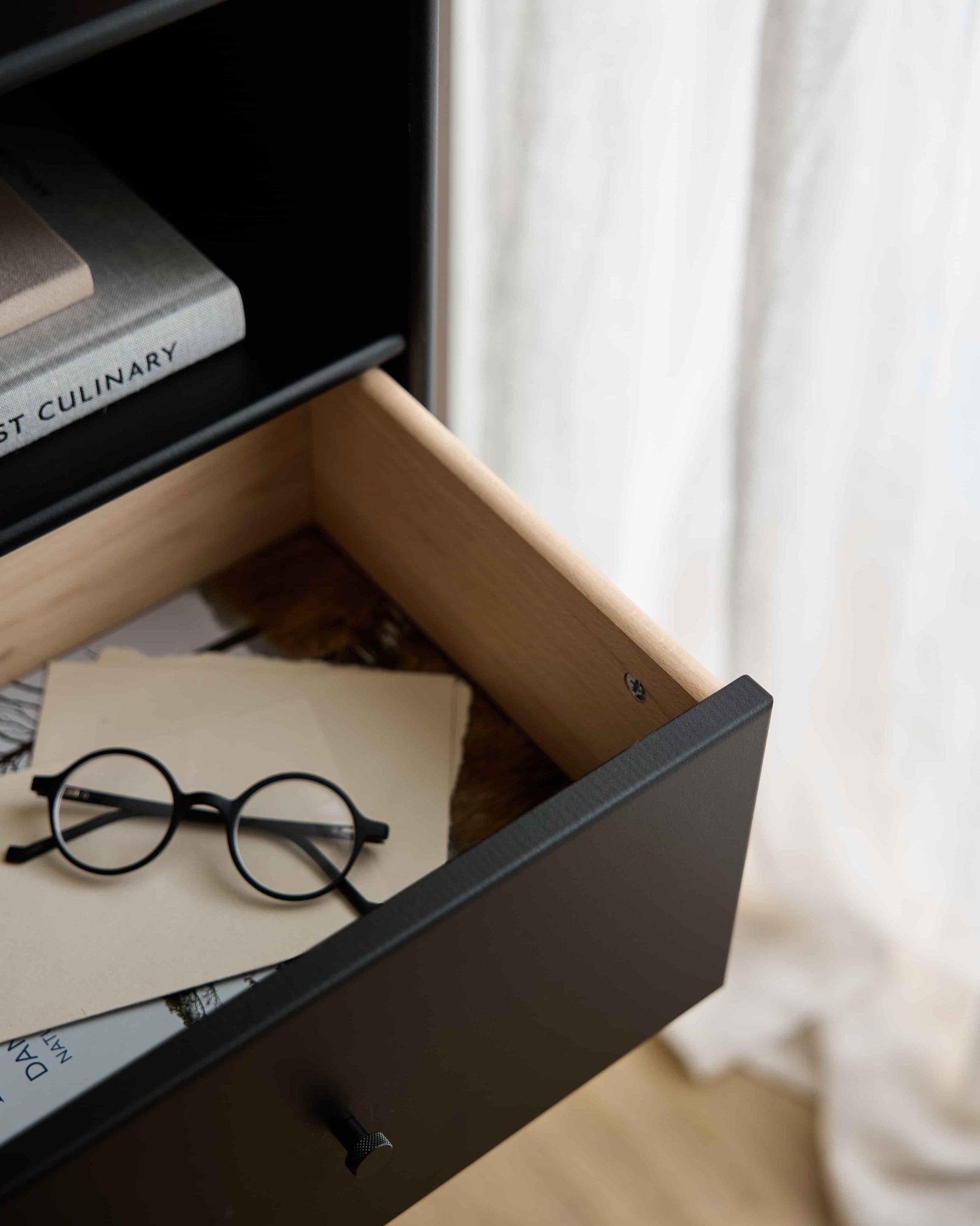Okrągłe czarne okulary i dokumenty w otwartej szufladzie regału WoodUpp Arka | Bookcase Drawer / Black, pod książkami, a delikatne zasłony miękko rozpraszają światło w tle.