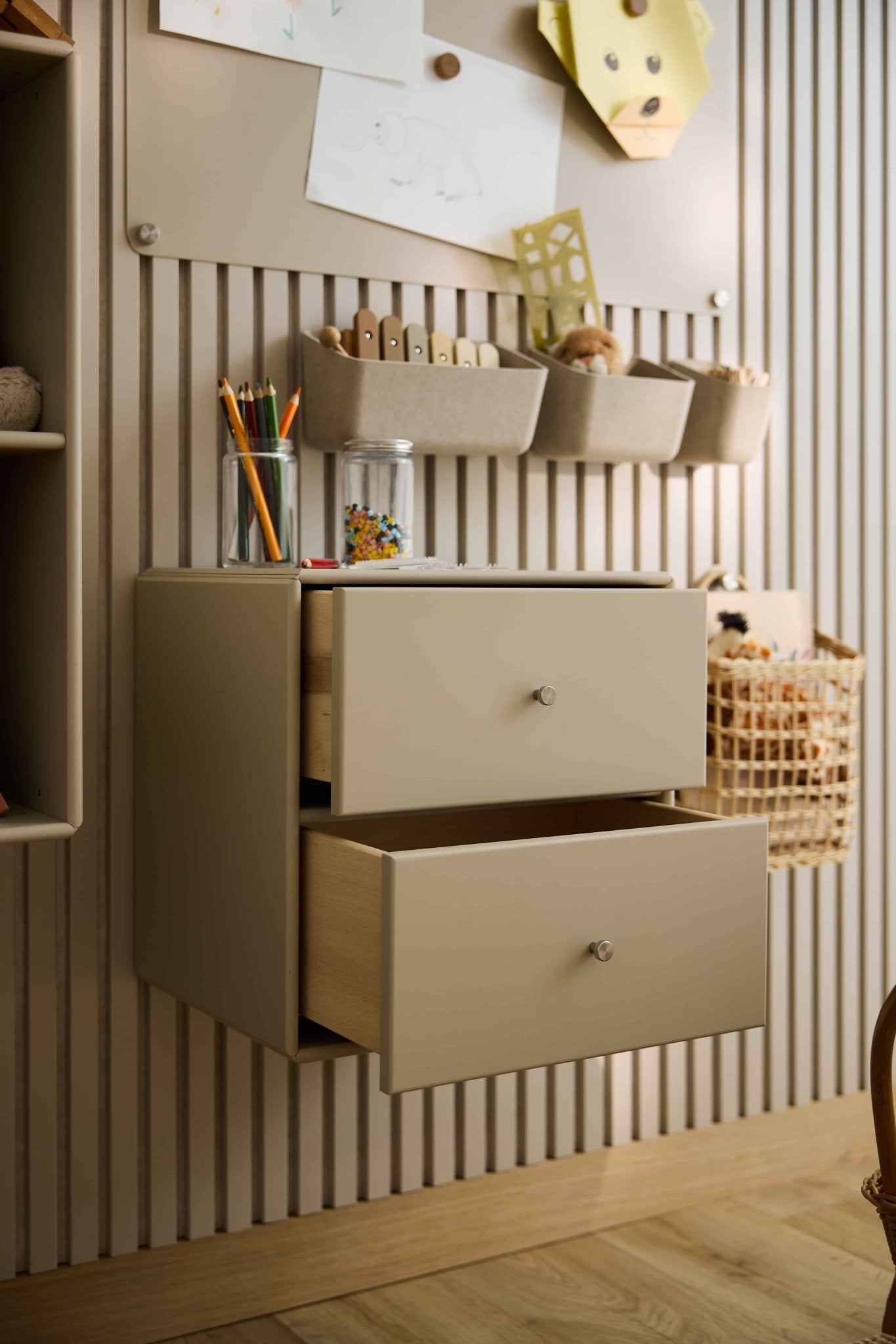 Dwie beżowe komody Arka Bookcase marki WoodUpp, częściowo otwarte, służą do przechowywania przyborów plastycznych i zabawek; w pobliżu znajdują się zamknięte szafki i kosze.