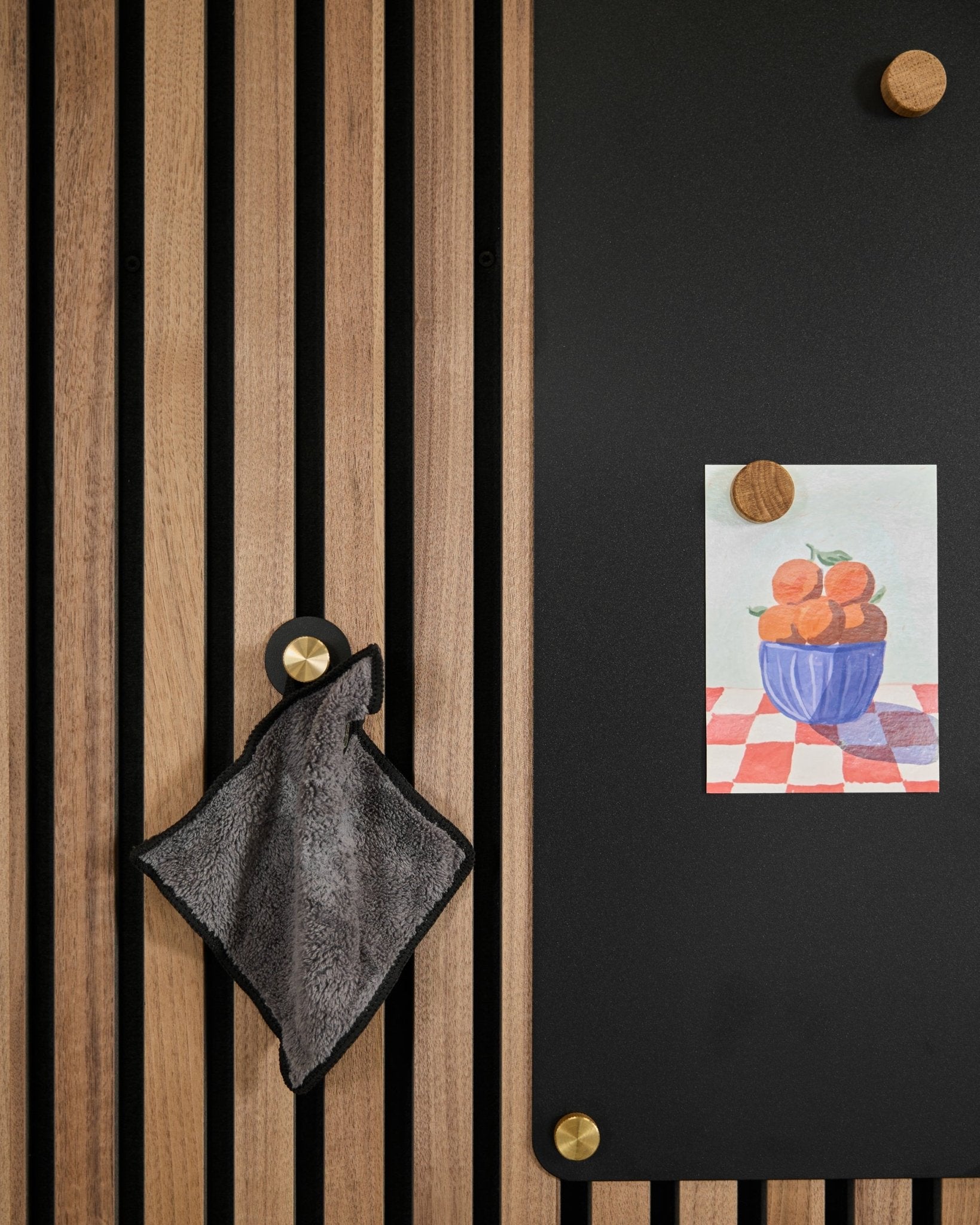 small ręcznik wisi na nordyckim panelu akustycznym z lamelami obok black tablicy z rysunkiem pomarańczy w misce, przymocowanym dwoma złotymi magnesami.