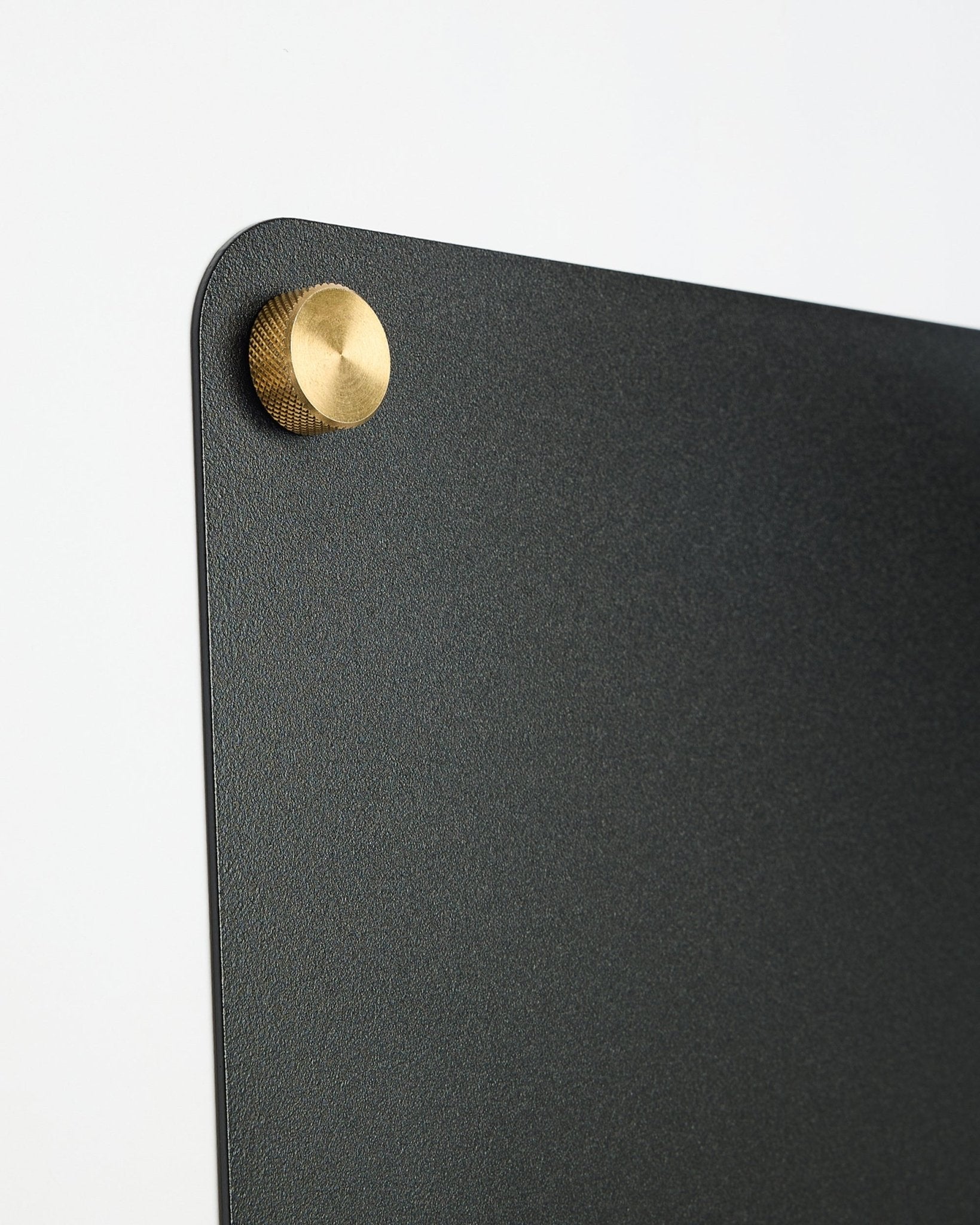 Zbliżenie na large black Norr tablicę magnetyczną od WoodUpp, część kolekcji CREATE, z okrągłym metalowym mocowaniem w kolorze złota-colored w górnym rogu.