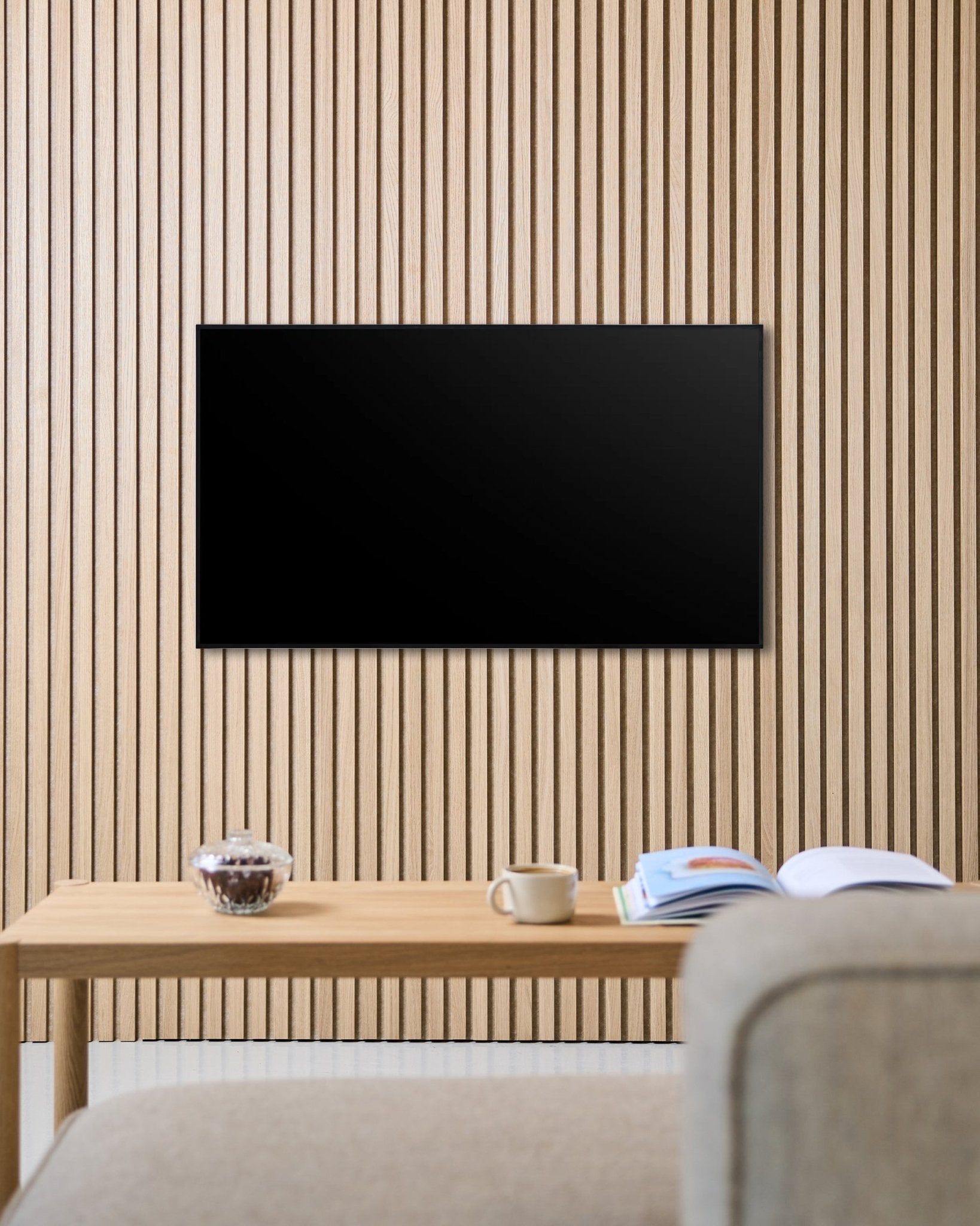 Uchwyt TV Fjell (small) od WoodUpp mocuje płaski ekran do nordyckiej ściany akustycznej z drewnianych lameli Akupanel, nad stołem z kubkiem, miską i otwartym magazynem obok sofy beige.