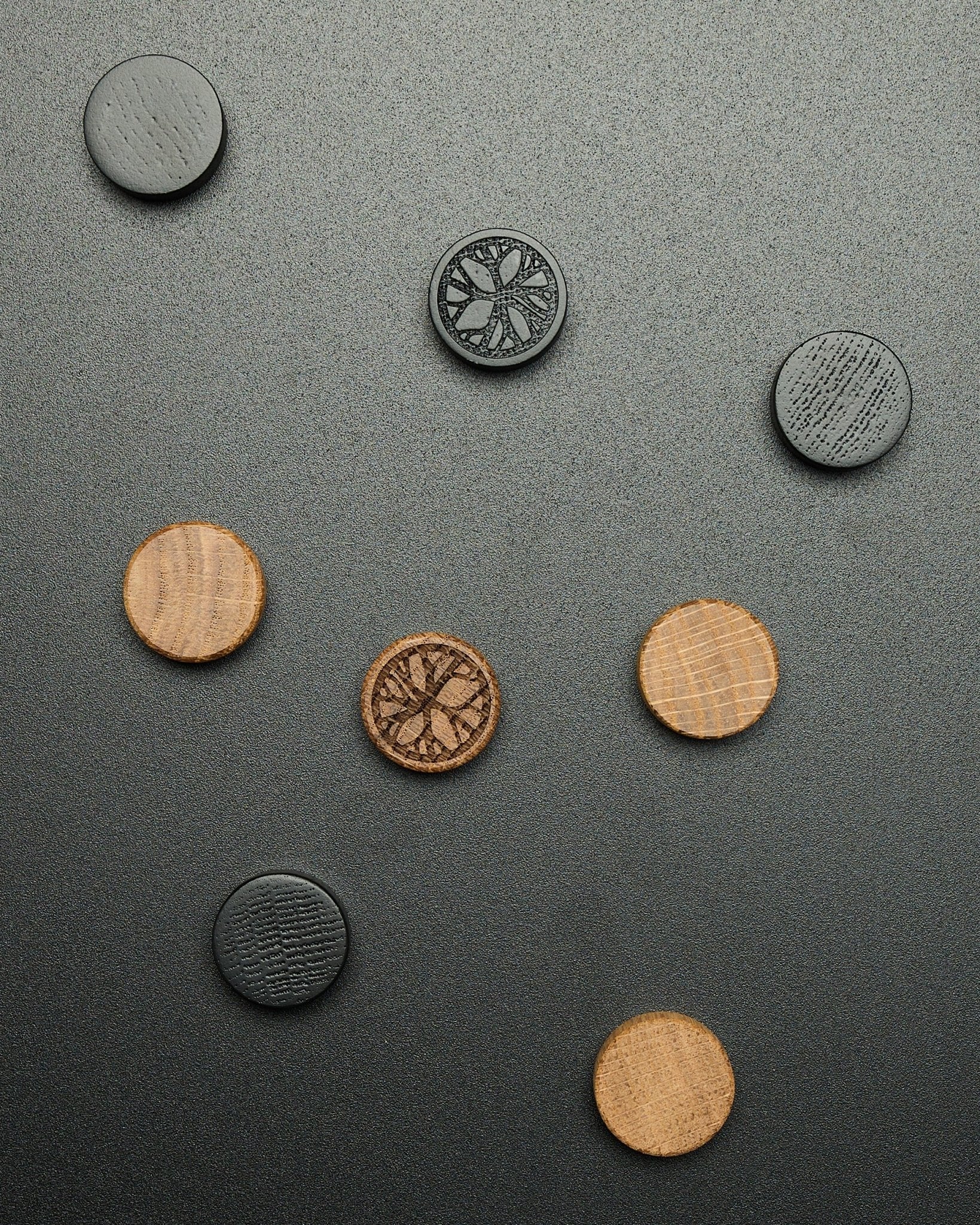 Cztery okrągłe magnesy black oak z zestawu WoodUpp Norr Magnet Set, luźno ułożone na ciemnej powierzchni o wyraźnej fakturze, zaprojektowane jako akcesoria Akupanel.