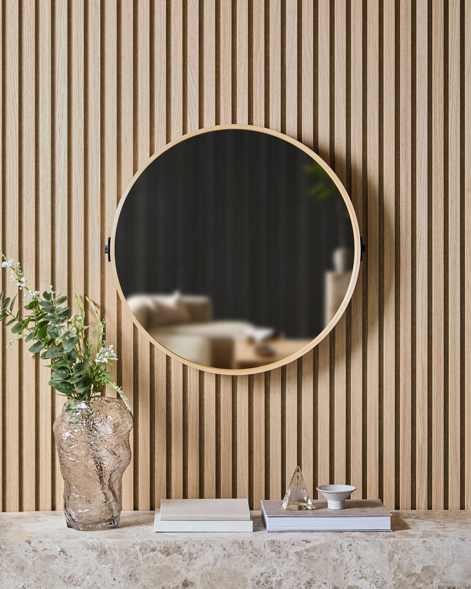 Linn | Mirror w kolorze Natural Oak od WoodUpp jest zamontowane na nordyckim panelu lamelowym Panel akustyczny nad kamienną półką z szklanym wazonem, książkami i dekoracjami.