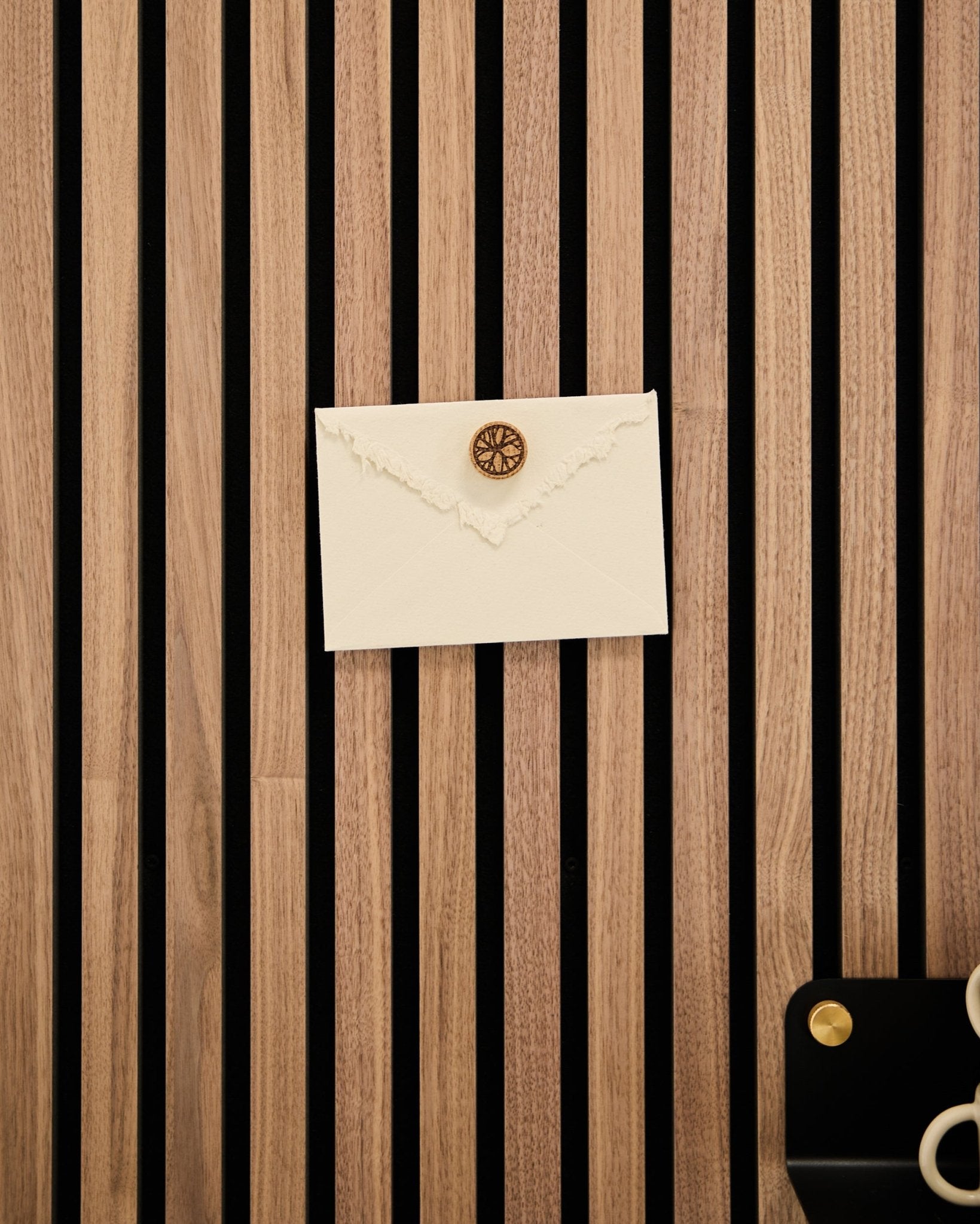 Kremowa koperta z lakiem jest przymocowana do dębowego akustycznego panelu lamelowego WoodUpp za pomocą zestawu Norr Poster Magnet w kolorze Natural Oak; obok znajdują się nożyczki i tacka.