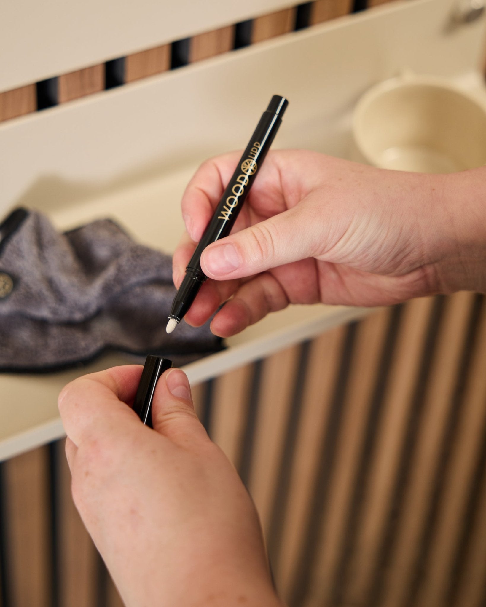 Osoba trzyma otwarty marker WoodUpp Norr | Blackboard set obok ściereczki z mikrofibry i kubka beige, produktów zaprojektowanych do użycia z akustycznymi panelami drewnianymi.