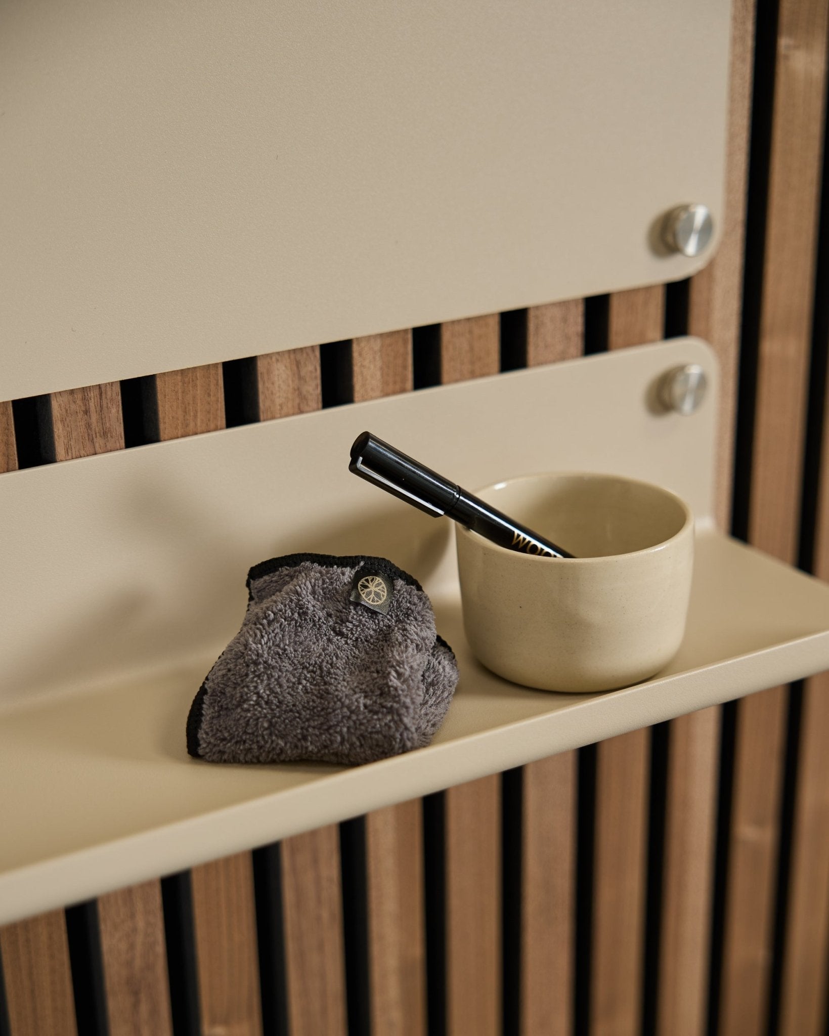 Zestaw Blackboard WoodUpp Norr zawiera kubek small beige, szczotkę z uchwytem black i ściereczkę do czyszczenia na półce beige z nordycką akustyczną boazerią drewnianą.