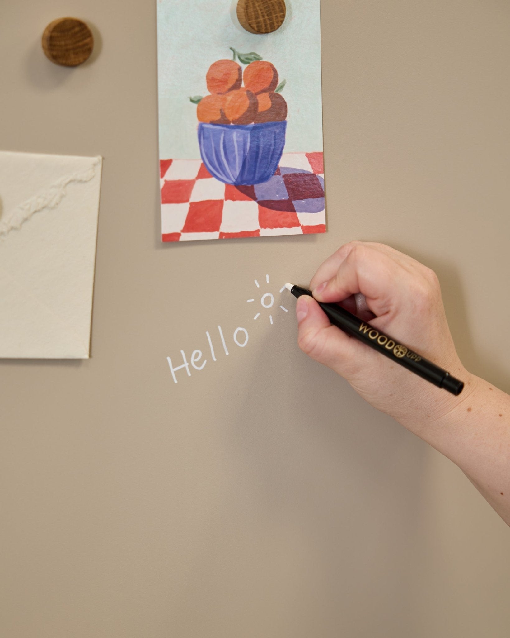 Dłoń pisze „hello” z rysunkiem słońca na tablicy Blackboard WoodUpp Norr, ustawionej obok pracy plastycznej przypiętej okrągłymi magnesami do Panel akustyczny.