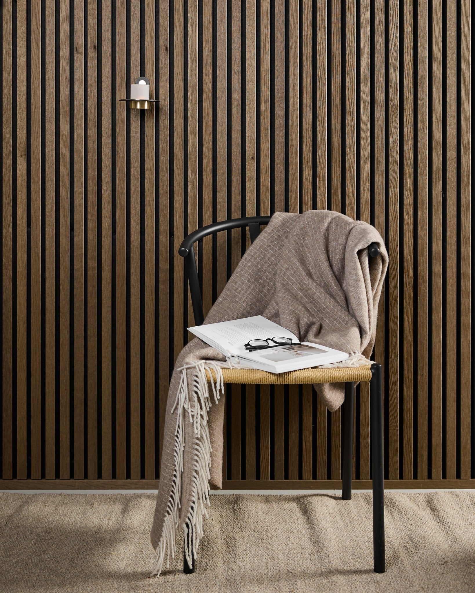 Nowoczesne krzesło z kocem beige, otwartą książką i okularami stoi obok akustycznego panelu drewnianego Akupanel oraz świecznika WoodUpp Norr w kolorach black i brass.