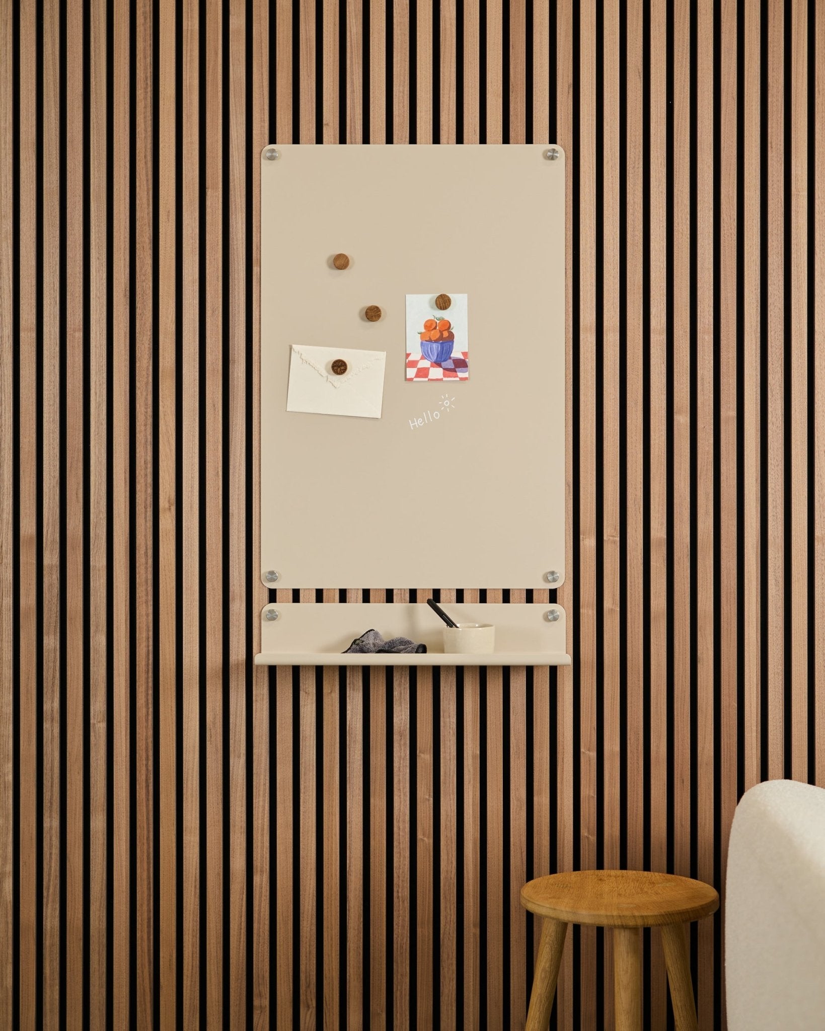 Tablica magnetyczna Beige Norr od WoodUpp zamontowana nad półką small na ścianie z paneli akustycznych z drewna Akupanel, obok stołka i białej sofy.