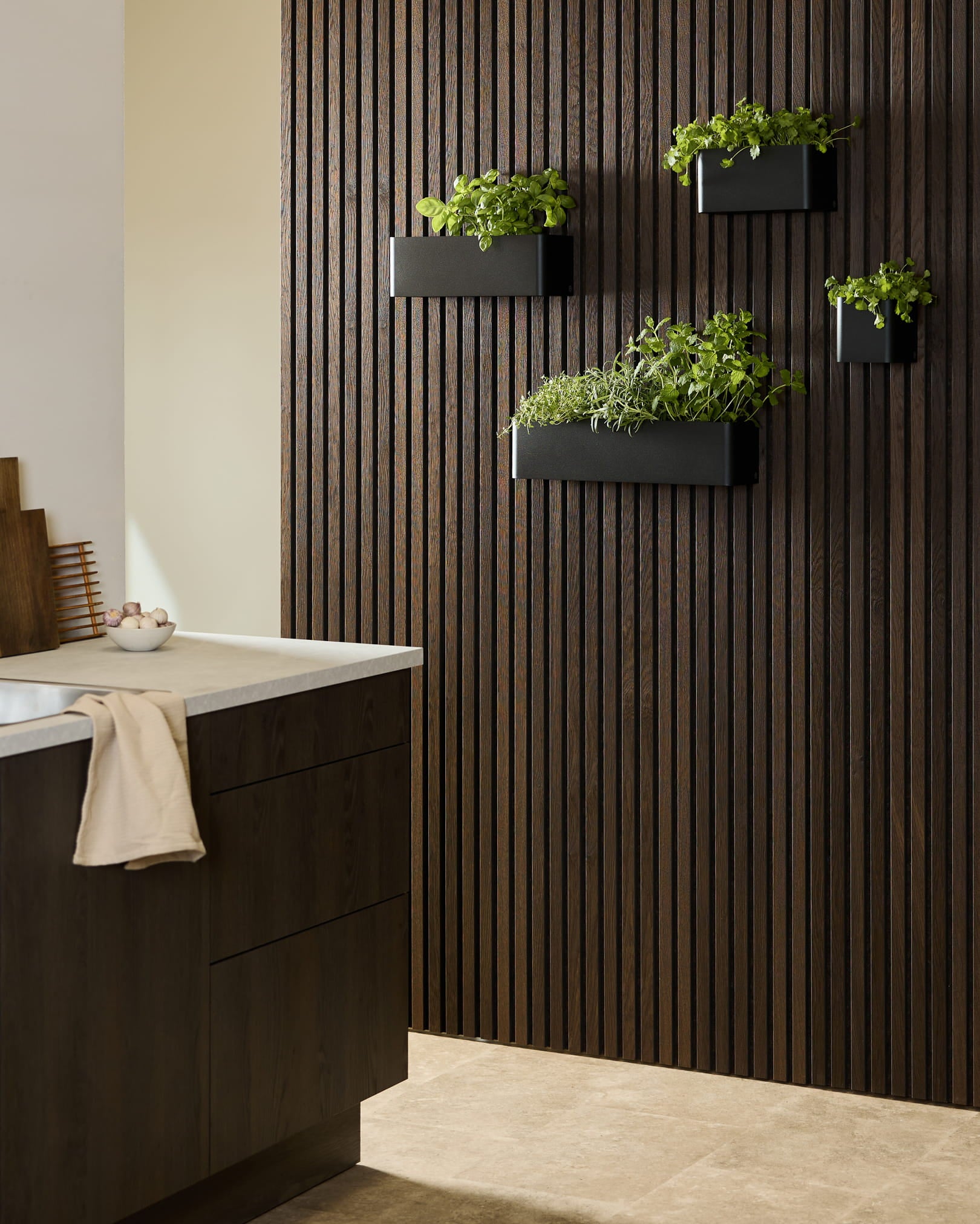Nowoczesna kuchnia z ciemnym drewnianym Panel akustyczny WoodUpp Akupanel, donicą Plant Box Norr black z ziołami oraz ciemną zabudową zwieńczoną białym blatem.