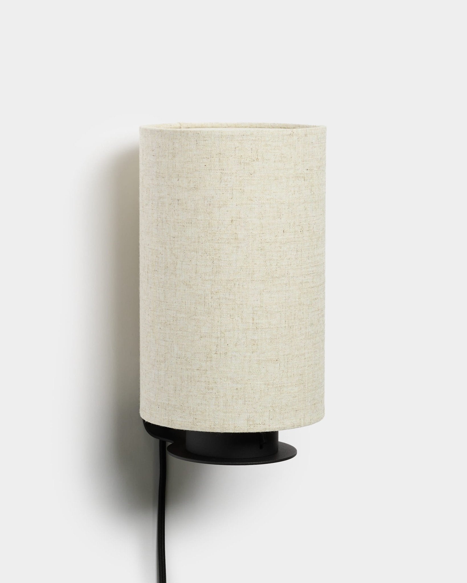Lampa ścienna Feya od WoodUpp z metalową podstawą w kolorze black, teksturowanym abażurem z mieszanki lnu i bawełny w kolorze beige oraz widocznym przewodem zasilającym w kolorze black. Nordycki, minimalistyczny design.