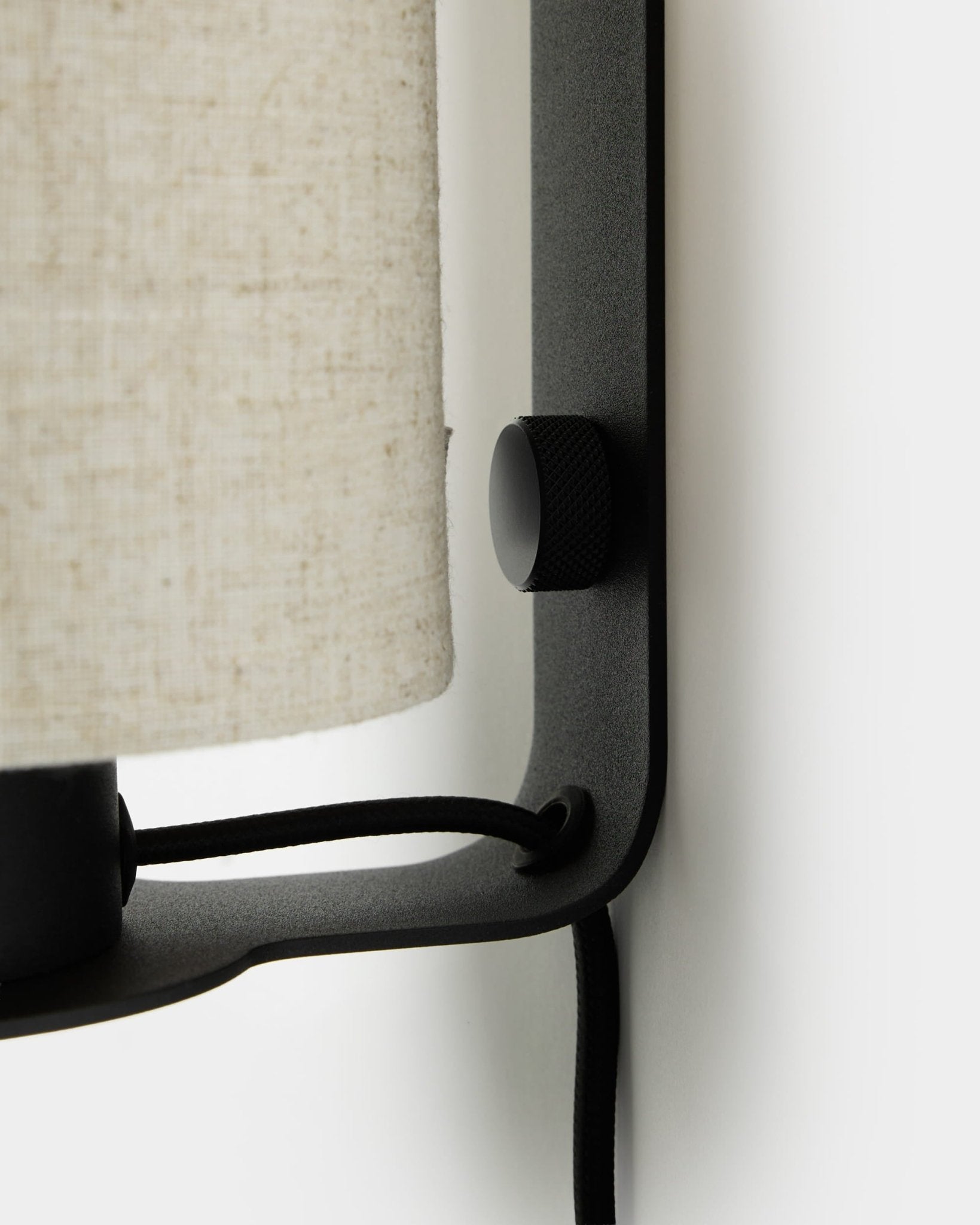 Zbliżenie na lampę ścienną Feya od WoodUpp w kolorach black i beige, z teksturowanym pokrętłem, abażurem z mieszanki lnu i bawełny oraz widocznym przewodem, zamontowaną na białej ścianie.
