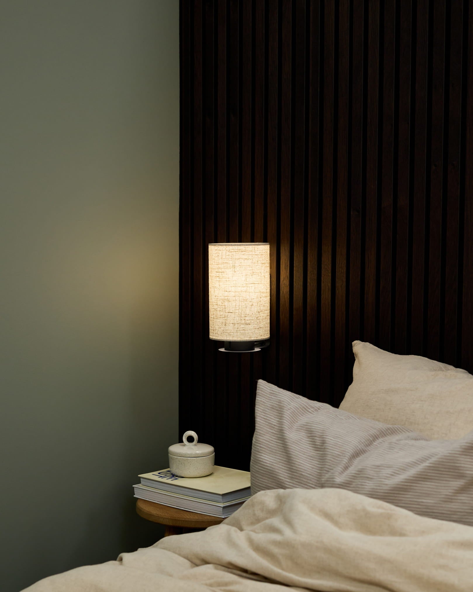 Kącik sypialniany z lampą ścienną Feya od WoodUpp w kolorach Black/Beige, pościelą w kolorze beige oraz stolikiem nocnym z książkami i ceramicznym kubkiem z pokrywką.