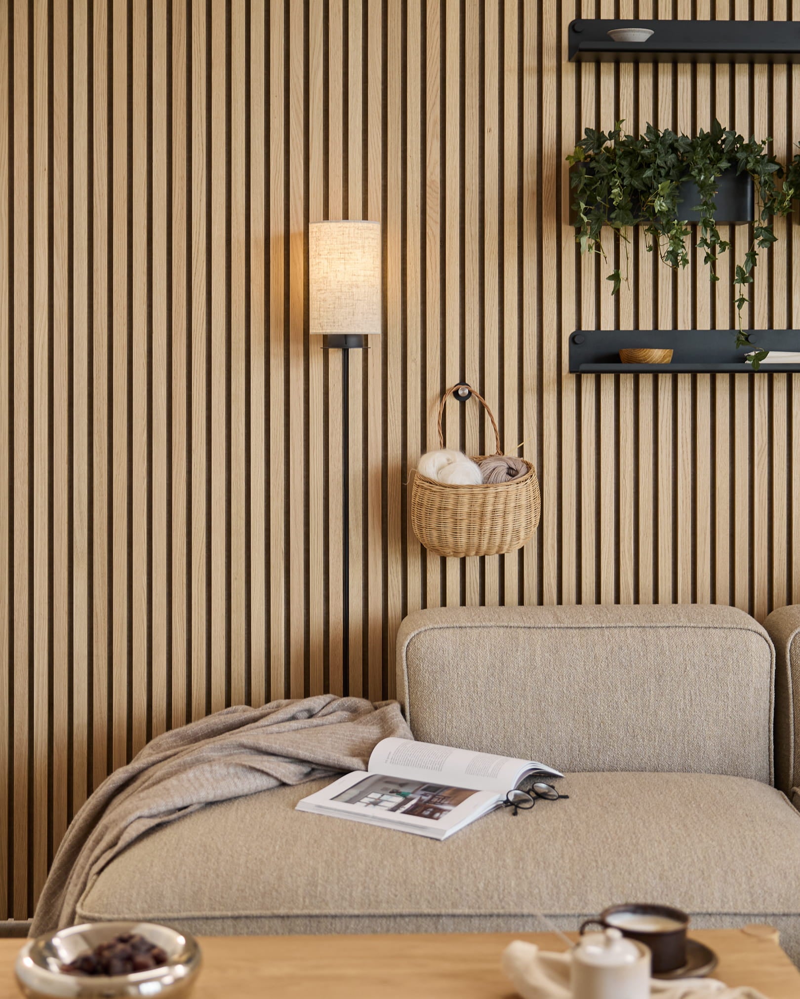 Sofa w kolorze Beige z kocem i książką obok akustycznej ściany lamelowej WoodUpp Akupanel, półki z roślinami i dekoracjami oraz lampa ścienna Feya w kolorach Black/Beige.