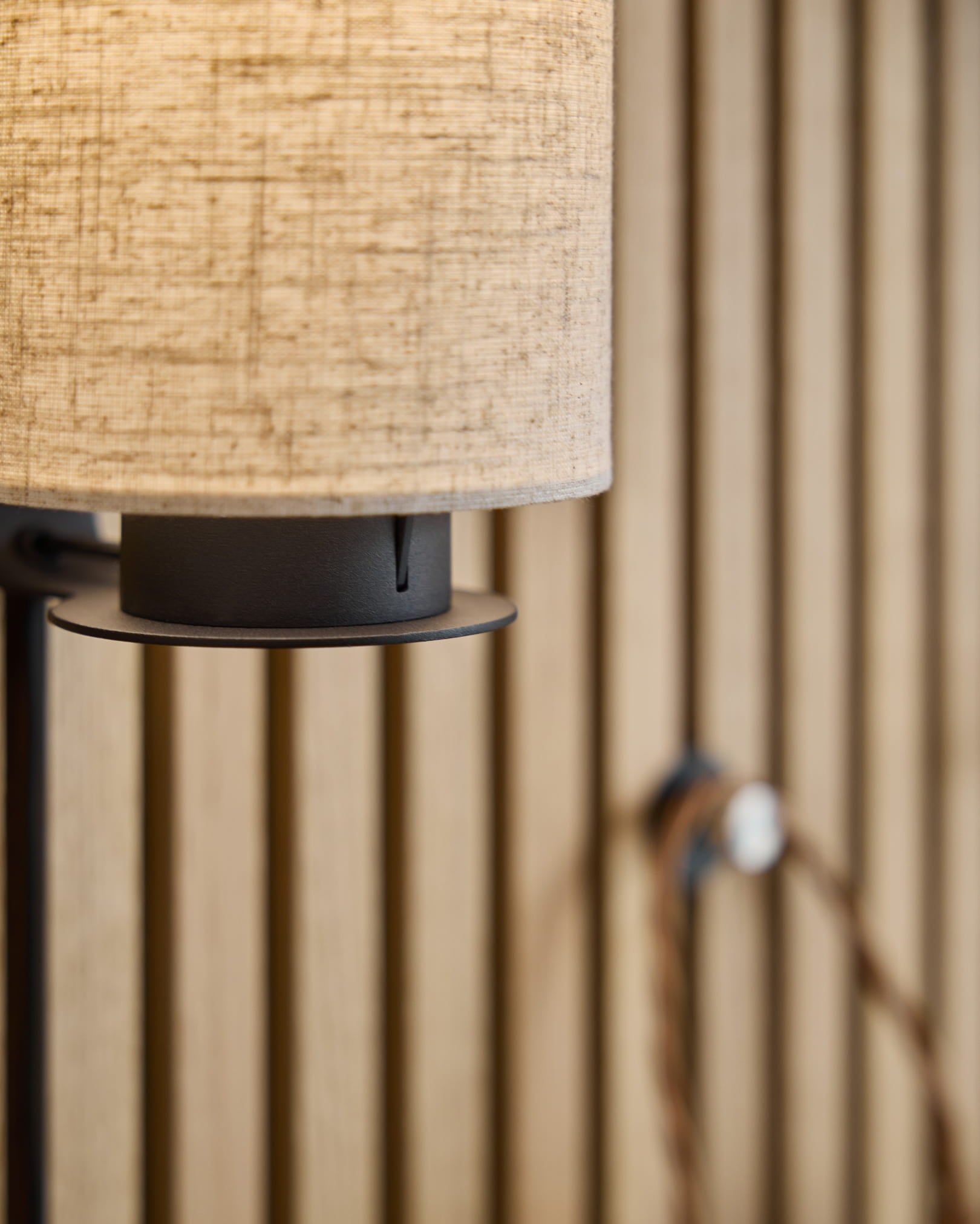 Lampa ścienna Feya / Black/Beige od WoodUpp z abażurem z mieszanki lnu i bawełny w kolorze beige, metalową podstawą w kolorze black i widocznym przewodem, zamontowana na nordyckim akustycznym panelu lamelowym.