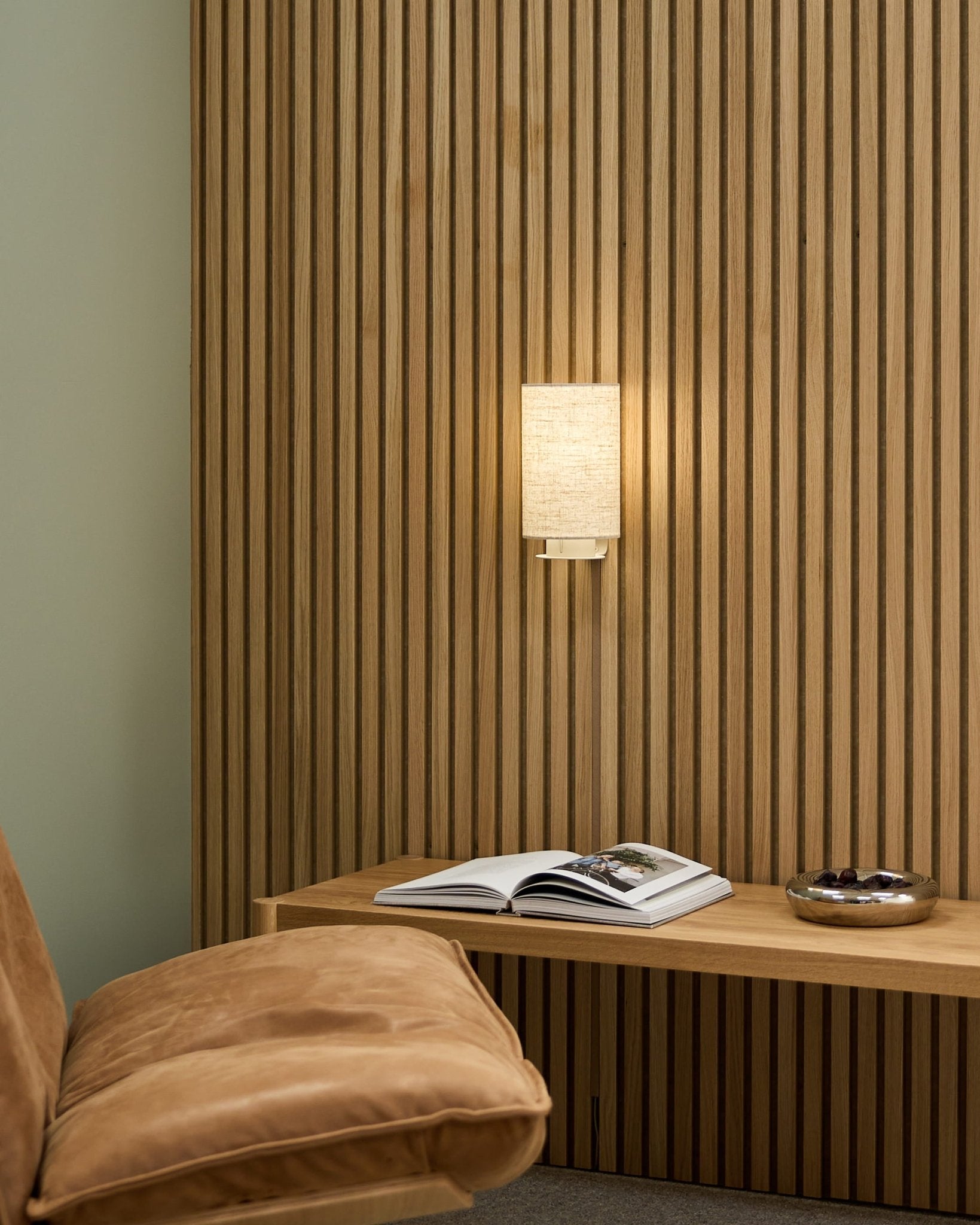 Minimalistyczne nordyckie wnętrze z krzesłem w odcieniu tan, akustyczną ścianą lamelową WoodUpp Akupanel, lampą ścienną Feya w kolorze Beige/Beige, otwartą książką na ławce i misą.