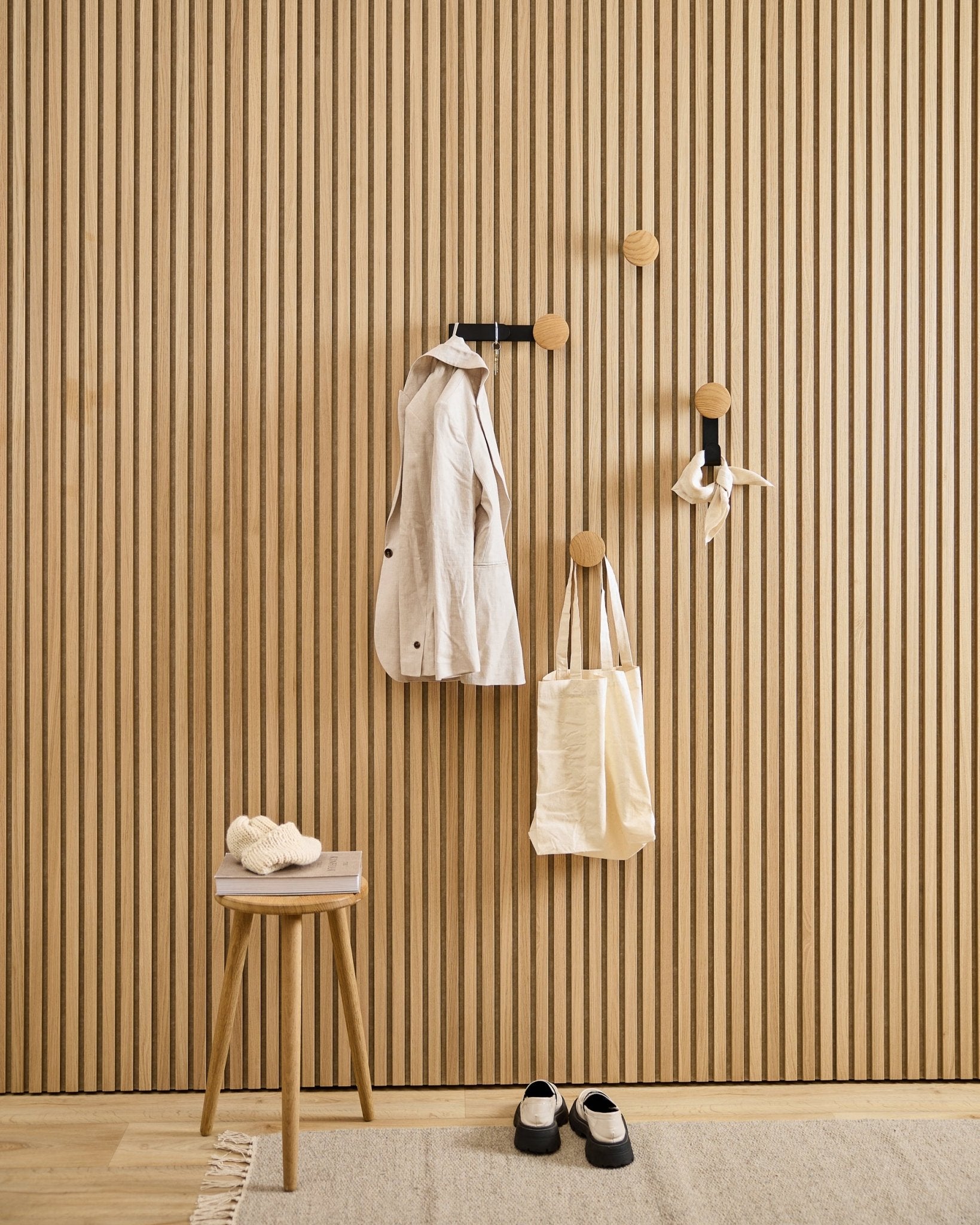 Minimalistyczny przedpokój z wieszakiem WoodUpp Linn Vertical Hanger w kolorze natural oak, z beige płaszczem, torbą typu tote, szalikiem, stołkiem z kapciami oraz butami w kolorze black i białym.