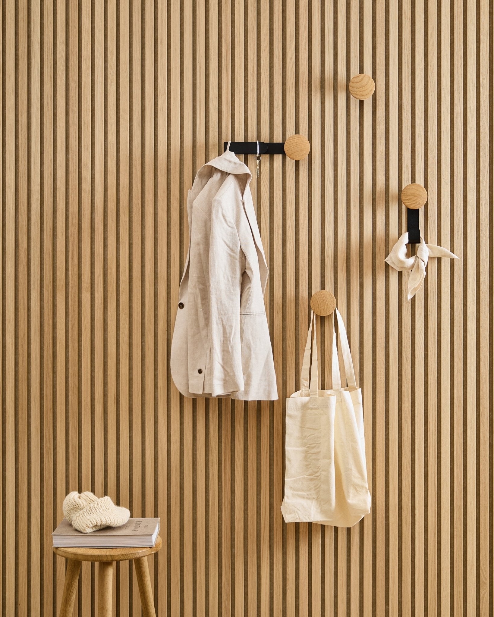 Kurtka w kolorze beige, torba tote i szalik wiszą na wieszaku WoodUpp Linn Horizontal w kolorze Natural Oak; stojący obok stołek podtrzymuje książkę i dzianinowy element.