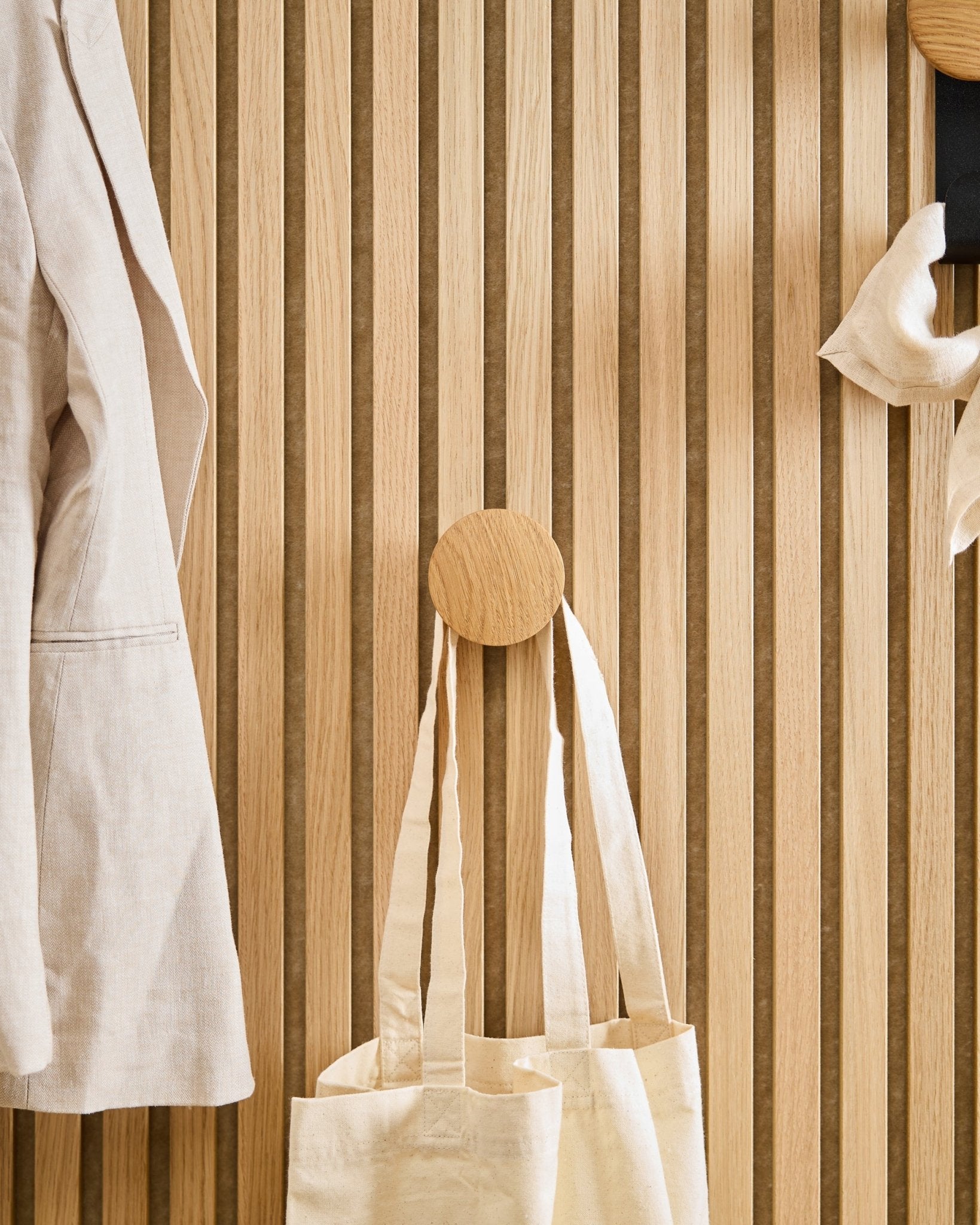 Na Linn I Single Hanger w kolorze Natural Oak od WoodUpp, zamontowanym na vertical Akupanel akustycznym panelu z drewnianych lameli, wiszą beige płaszcz i torba tote.