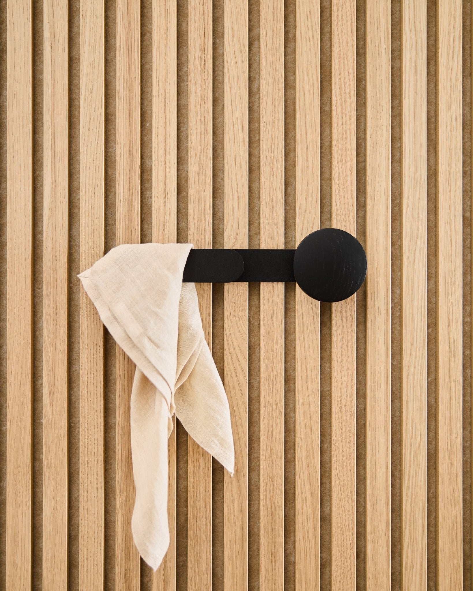Tkanina w odcieniu beige swobodnie opada na wieszak WoodUpp Linn Horizontal w kolorze Black Oak, zamocowany do akustycznego panelu ściennego z lamelami w odcieniu vertical.