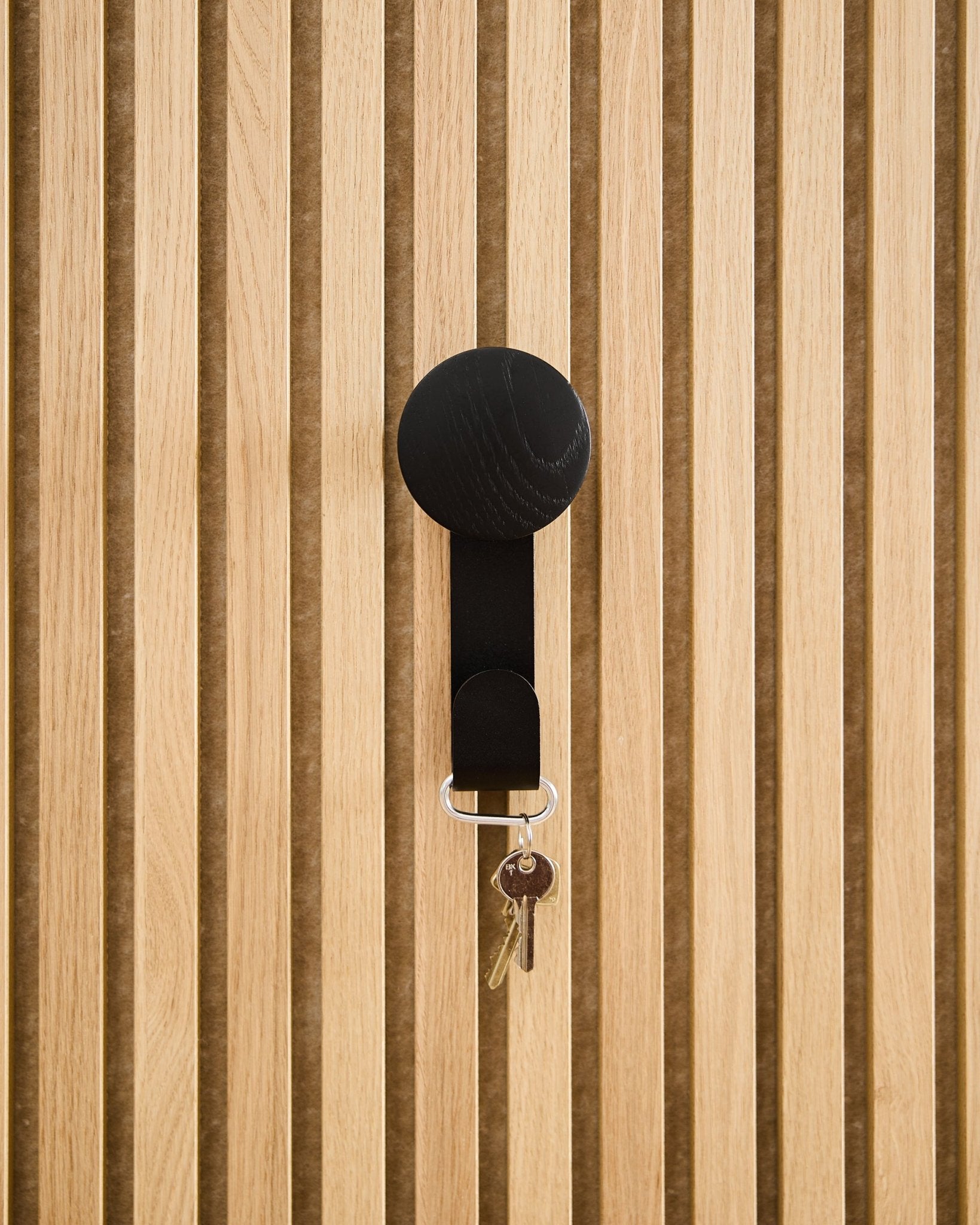 Wieszak Linn Vertical w kolorze Black Oak od WoodUpp utrzymuje brelok z dwoma kluczami i okrągłą zawieszką, zamocowany na teksturowanym akustycznym panelu drewnianym Akupanel.