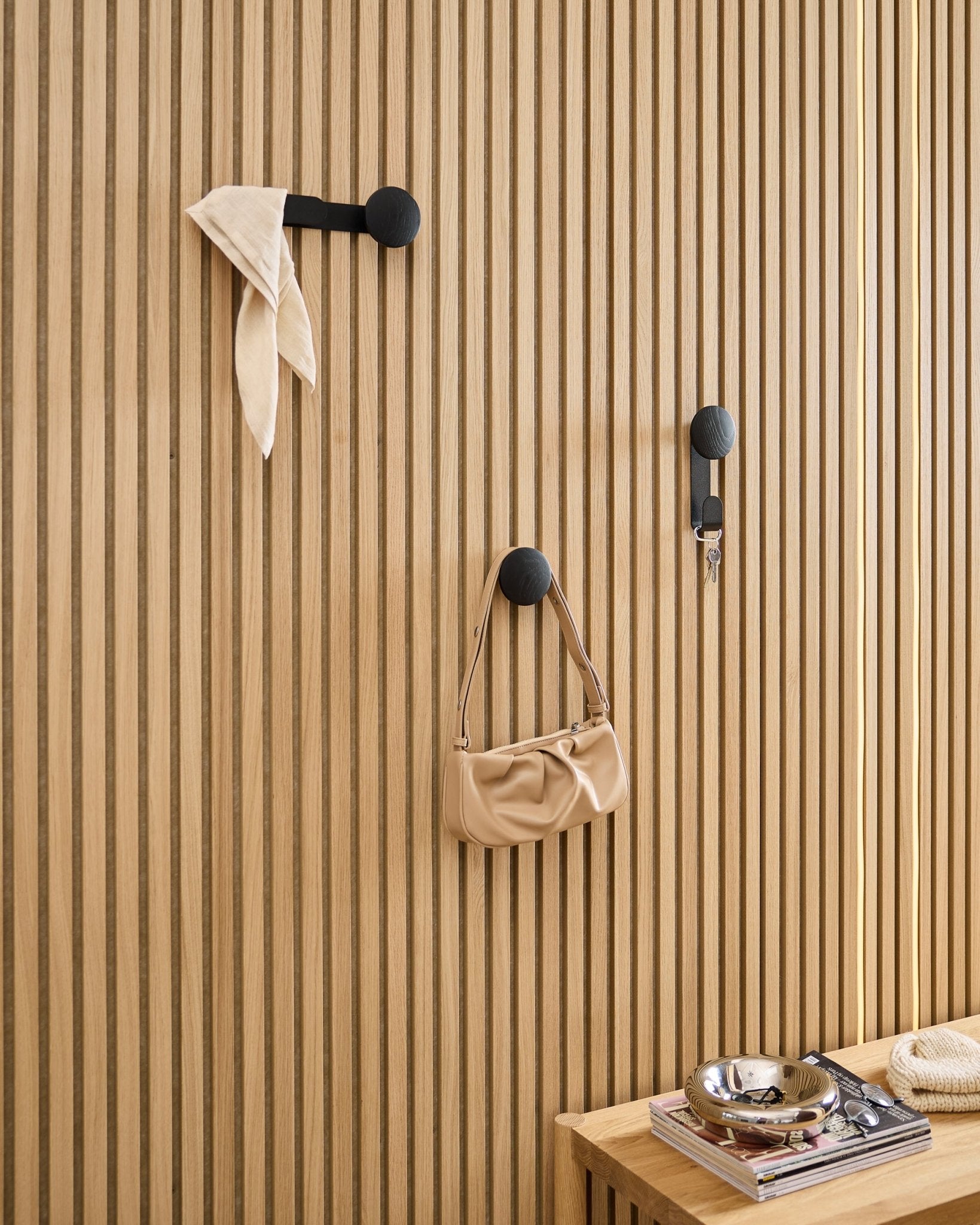 WoodUpp Linn Single Hanger w kolorze Black Oak utrzymuje szalik, torbę beige i klucze na panelu akustycznym z drewna black Akupanel; obok stoi ławka z książkami i metalową misą.