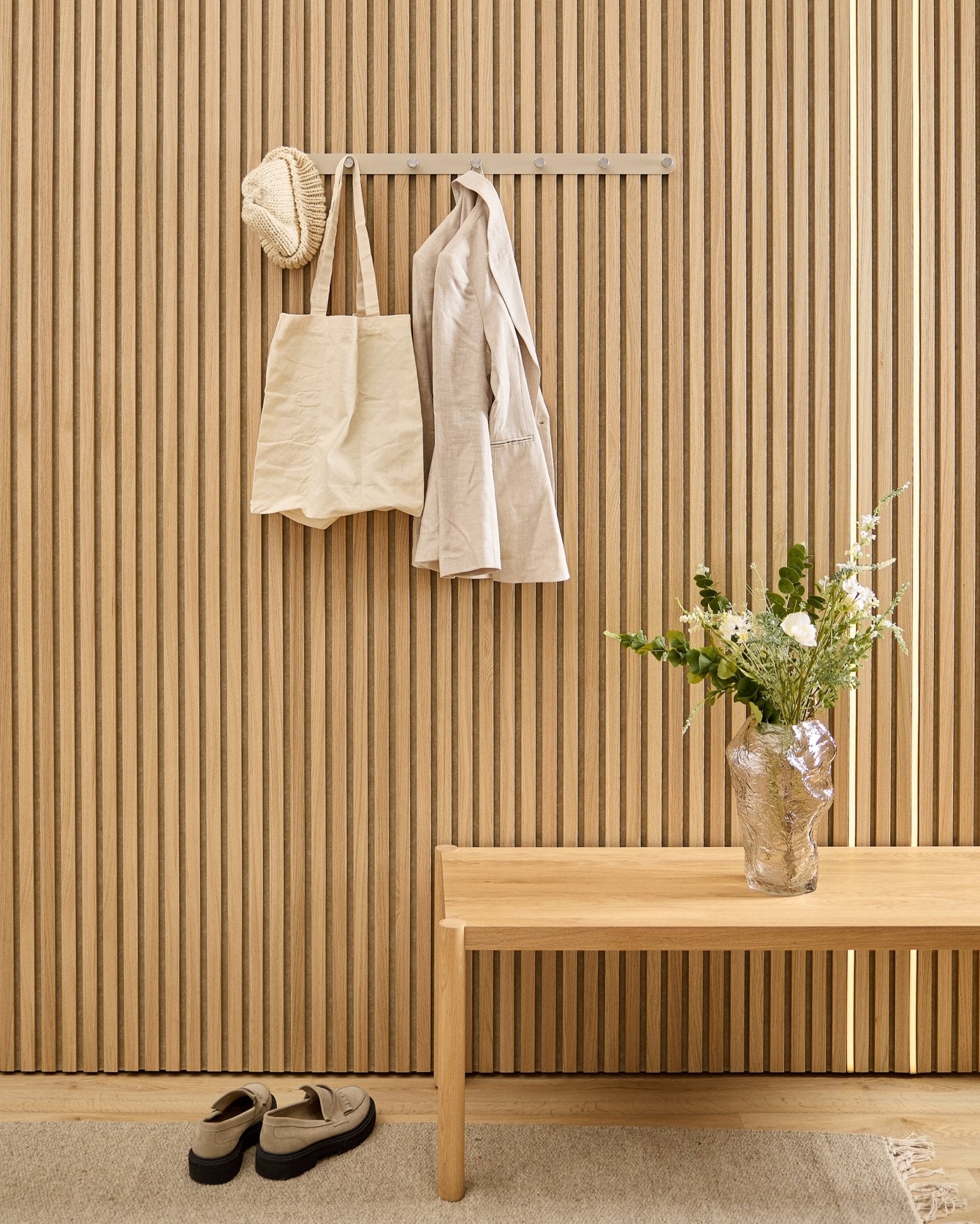 Na WoodUpp Norr Coat Hook Rack wiszą beige torba, czapka i koszula, nad butami; obok stoją ławka i wazon przy akustycznym panelu ściennym z listew w pasy.