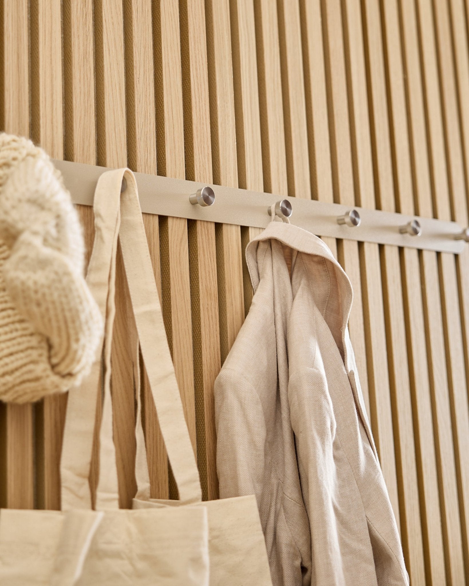 Na WoodUpp Norr Coat Hook Rack (Beige/stal nierdzewna Steel, X7), zamocowanym na jasnym drewnianym panelu ściennym z listew Akupanel, wiszą beige torba tote, dzianinowa czapka i lniana koszula.