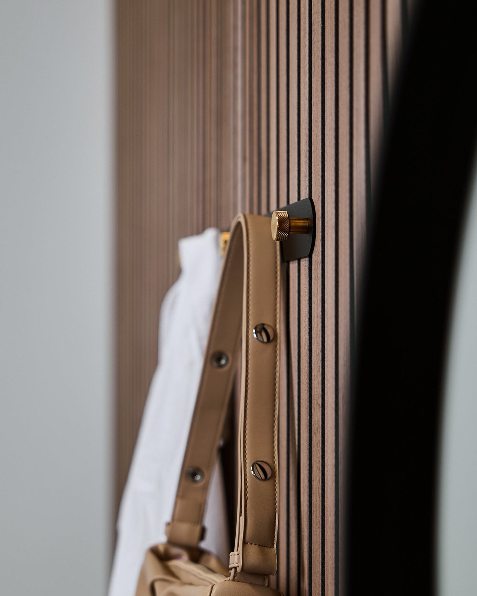 Jasnobrązowa torba i biała koszula wiszą na wieszaku ściennym WoodUpp Norr (Black/Brass, X5), zamontowanym na akustycznym panelu lamelowym vertical Akupanel.