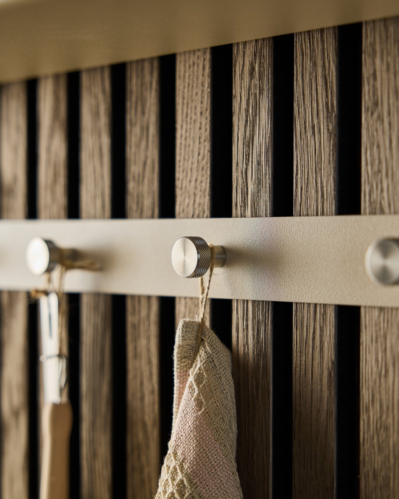 Zbliżenie na wieszak ścienny WoodUpp Norr (Beige/stal nierdzewna Steel, X5) zamontowany na drewnianej ścianie lamelowej Panel akustyczny, na którym wisi ręcznik i narzędzie z uchwytem.
